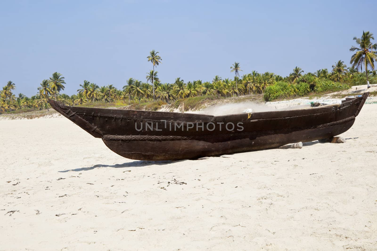 Horizontal landscape of a Goan fishermans wooden boat with his fishing nets and tackle on board resting on ramps on a white sandy beach with a coconut palm tree lined shoreline.