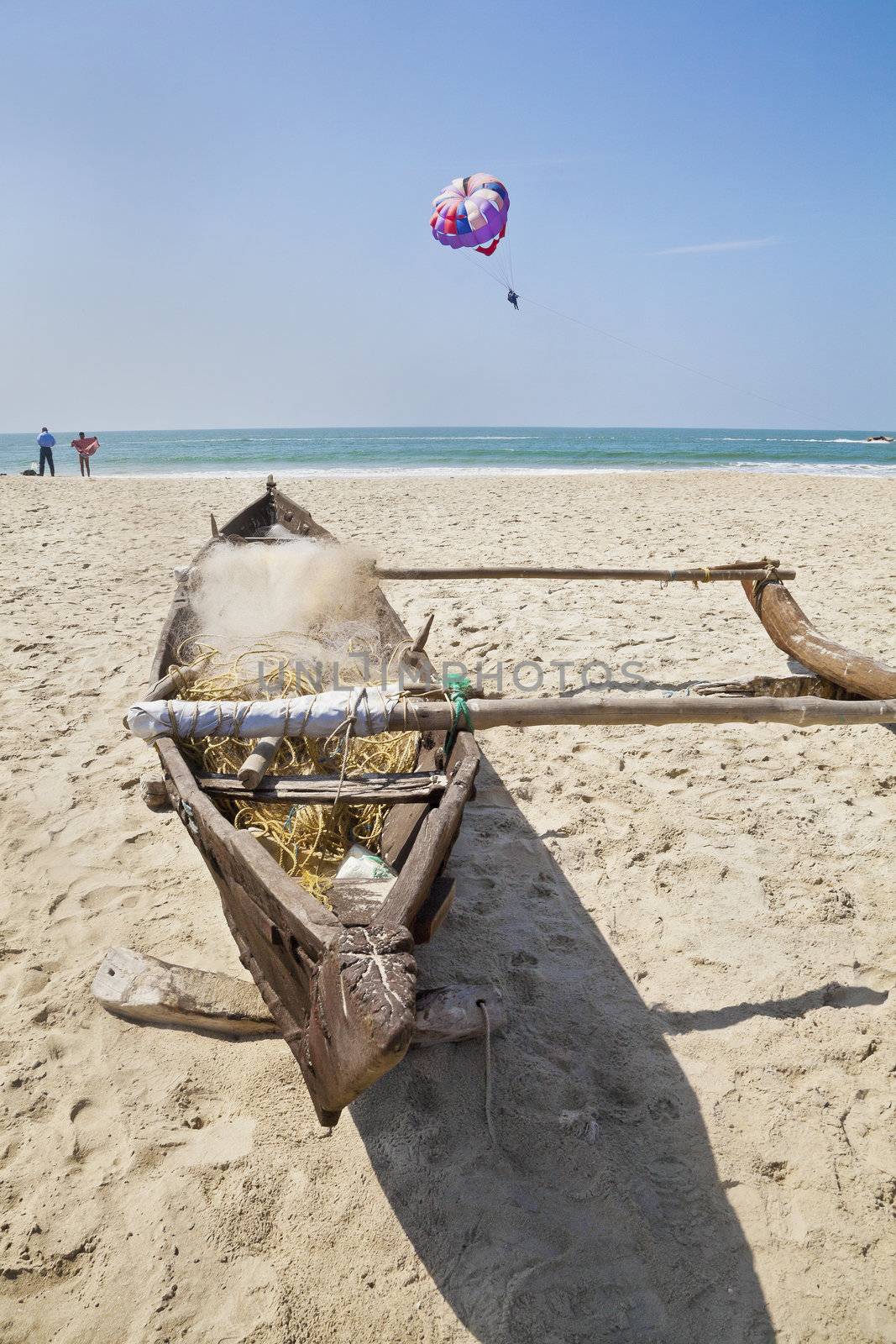 Horizontal landscape of a Goan beach in India with a moored fishermans boat on a sandy beach, bathers about to undress an get in the ocean while a paraglider drifts by