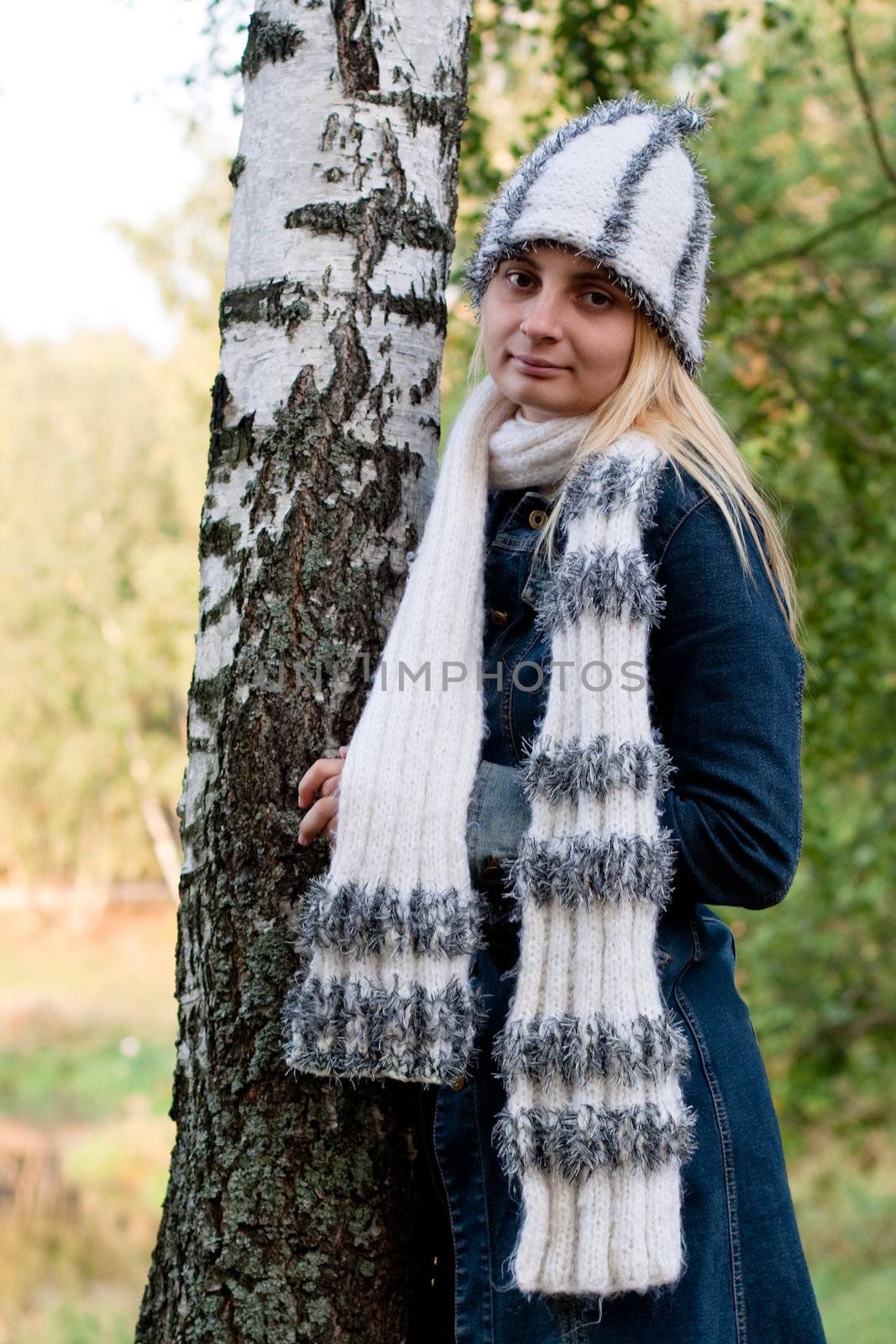 A standing girl in a cup and scarf and jeans coat in a forest
