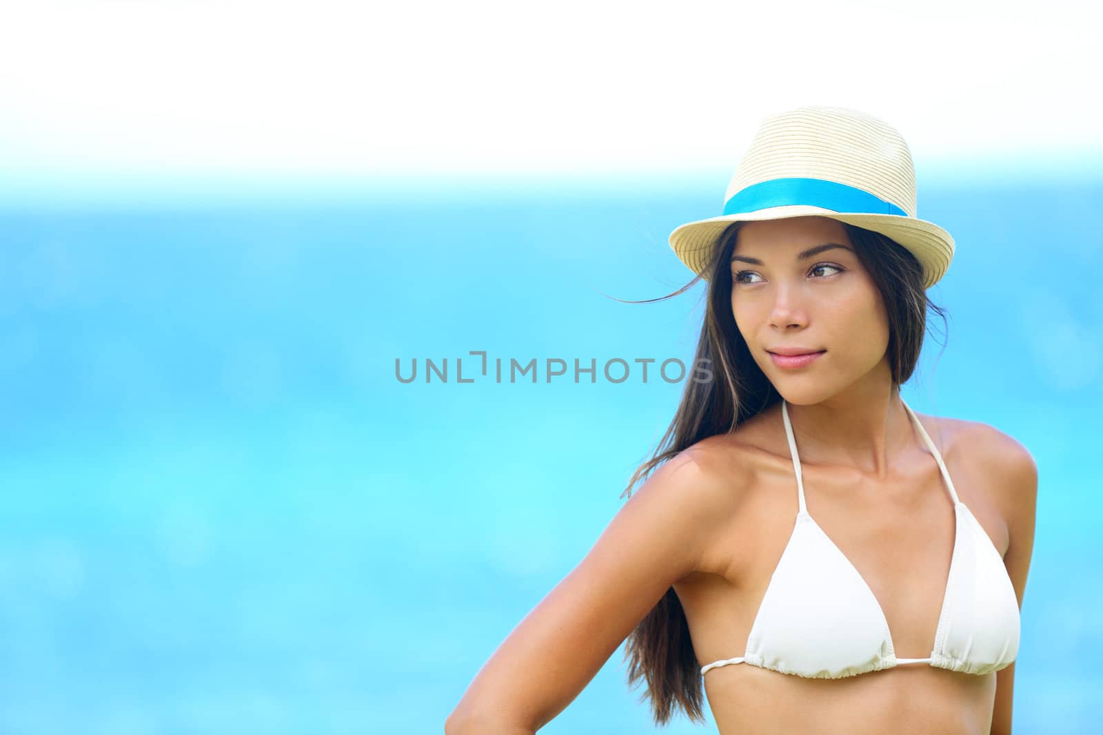 Woman beach portrait looking to side with wearing hat and beachwear bikini. Beautiful young modern multiracial female model looking serious with subtle smile. Half asian and half caucasian model outdoors.
