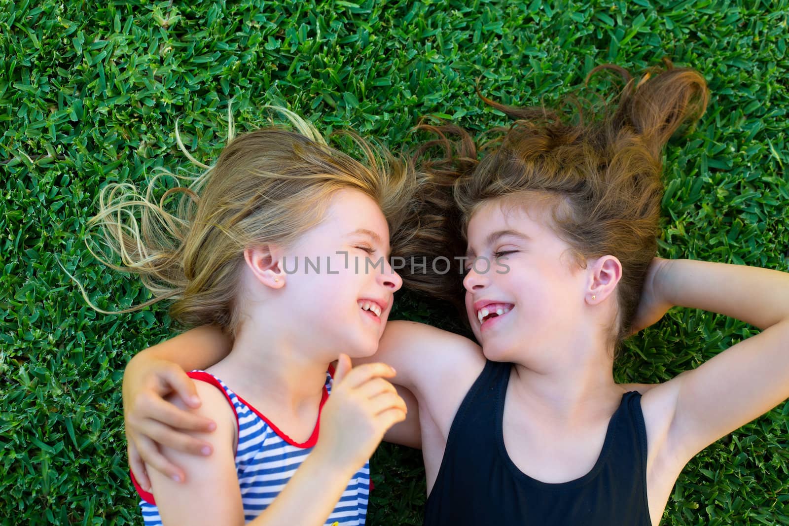 Blond and brunette sisters kid girls smiling lying on garden grass