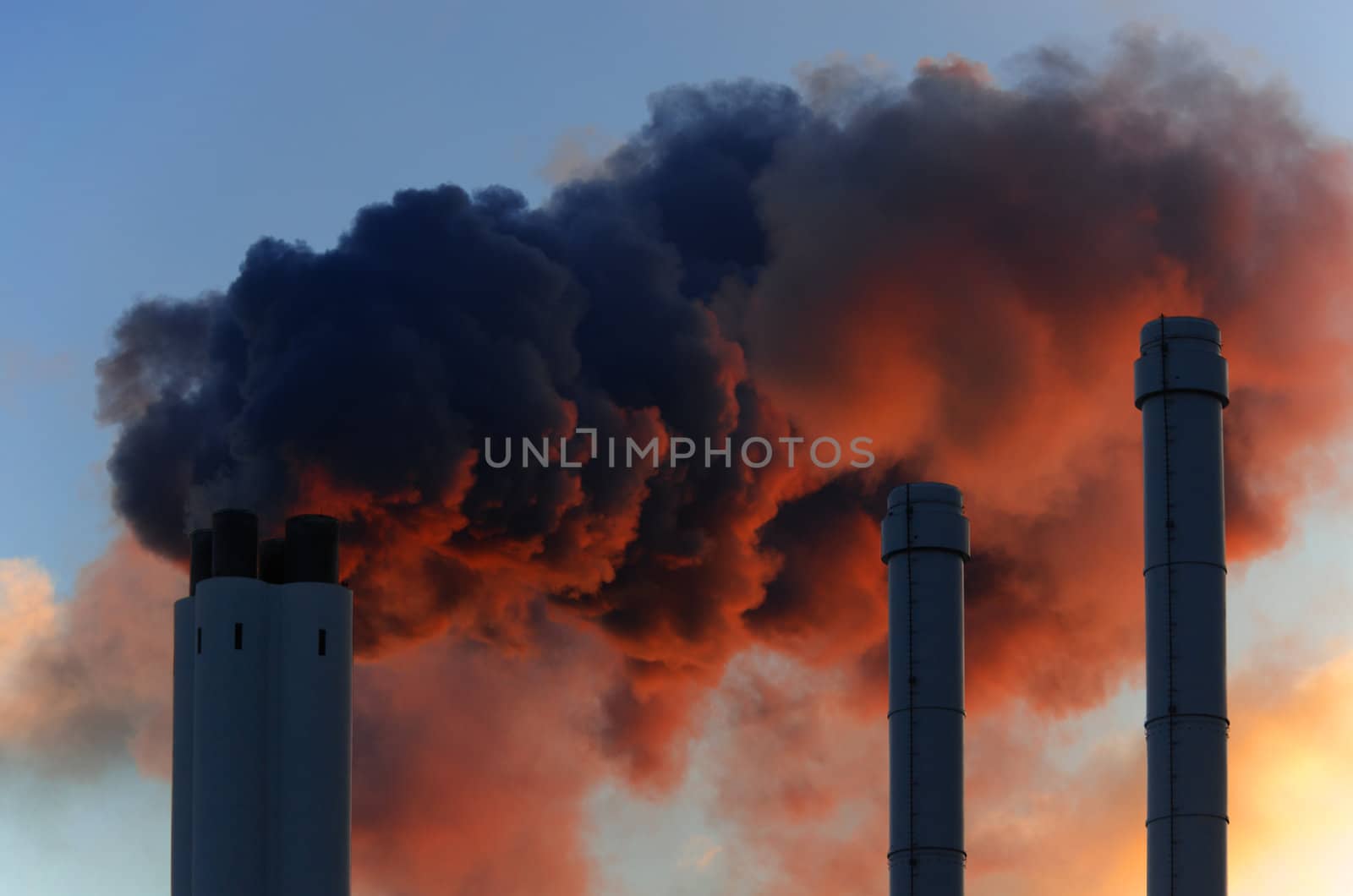 Pollution concept of smoking chimneys. The setting sun illuminates the vapor from below, giving it an ominous impression, like that of volcano smoke.