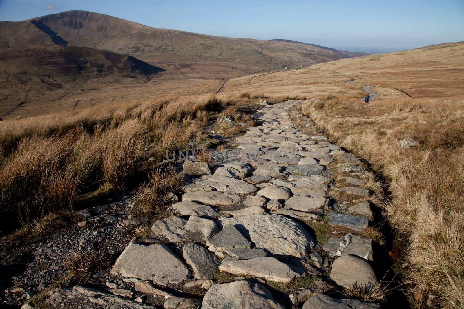 A well maintained stone made footpath with a grassy landscape leading towards hills.