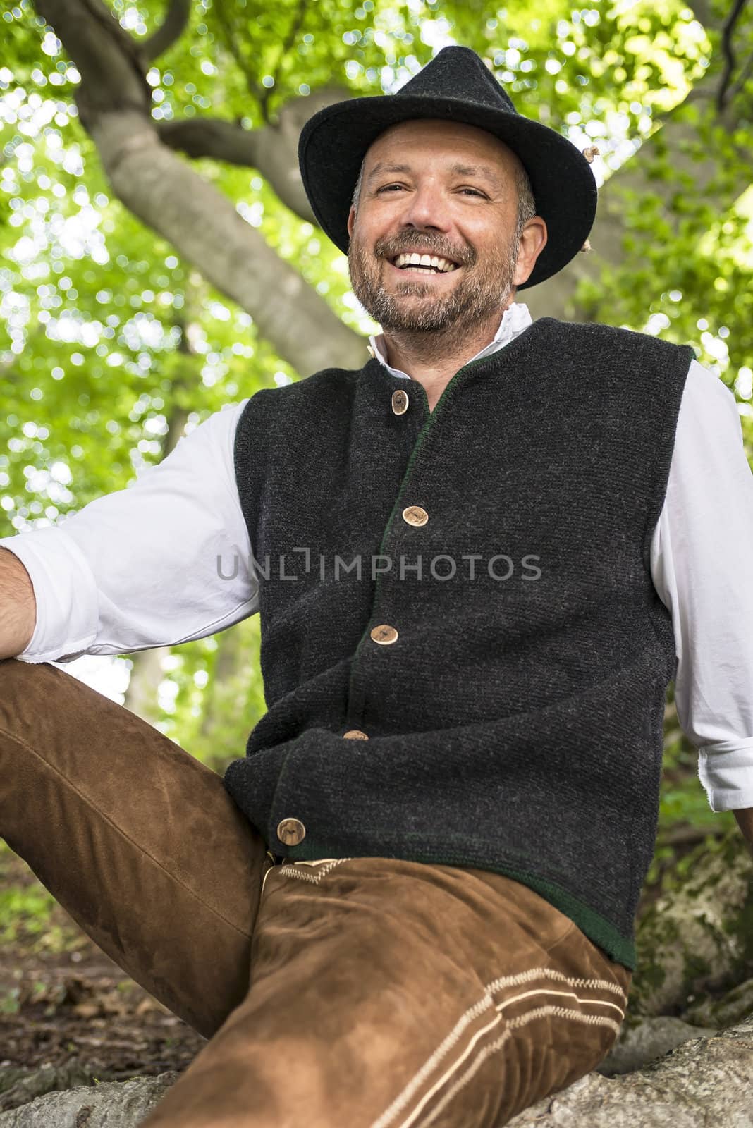 Laughing man in Bavarian costume sitting in a forest on a sunny day