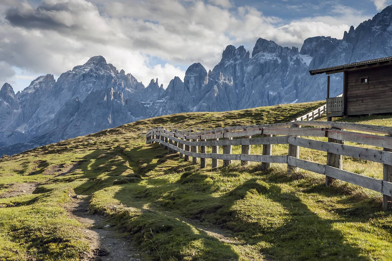 Evening scenery in South Tirol with green meadows, cottage, fence, alps and clouds on blue sky