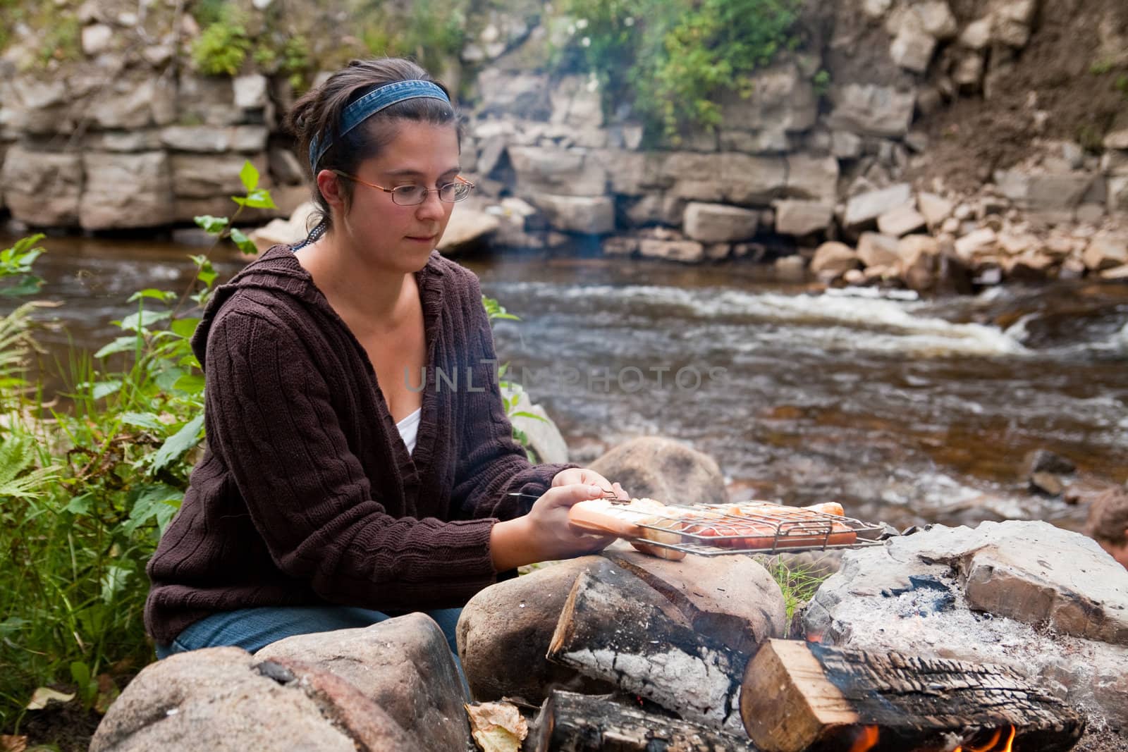Woman cooking hotdogs over a campfire near a river