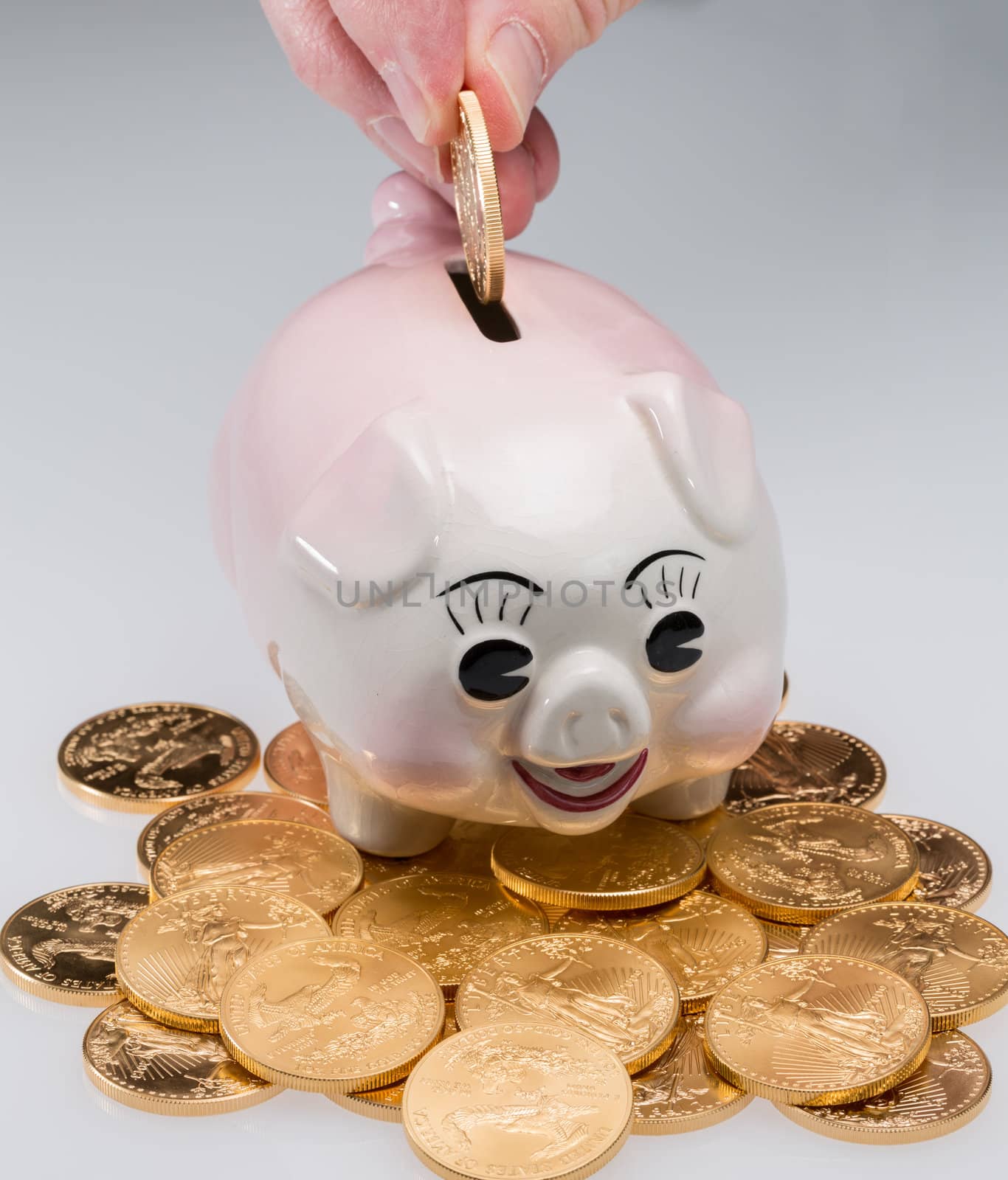 Fingers placing a single gold coin into the slot on a piggy bank with stack of coins around pig
