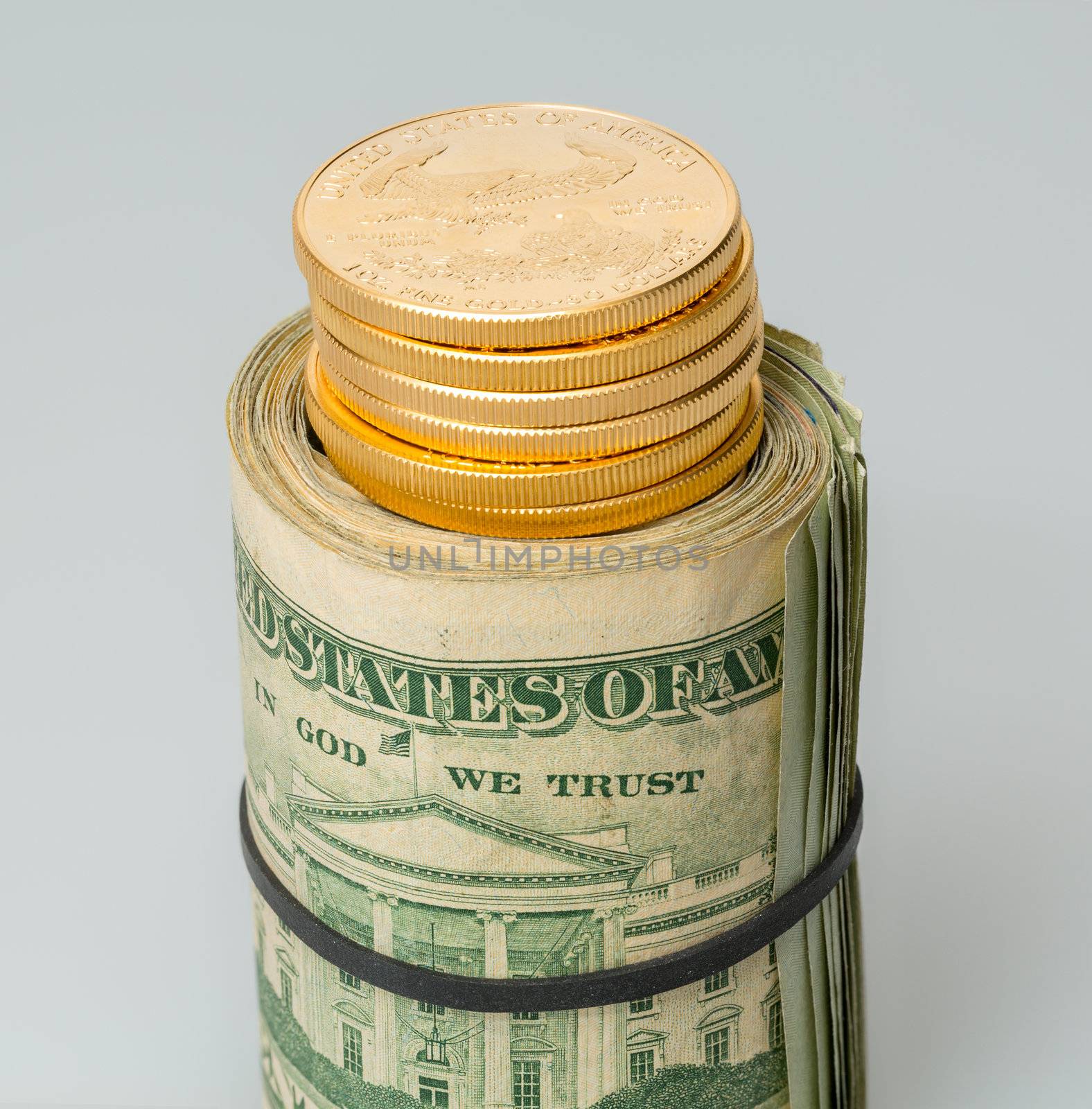 Gold coins standing on roll of many $20 dollar bills and notes secured by elastic band on white table