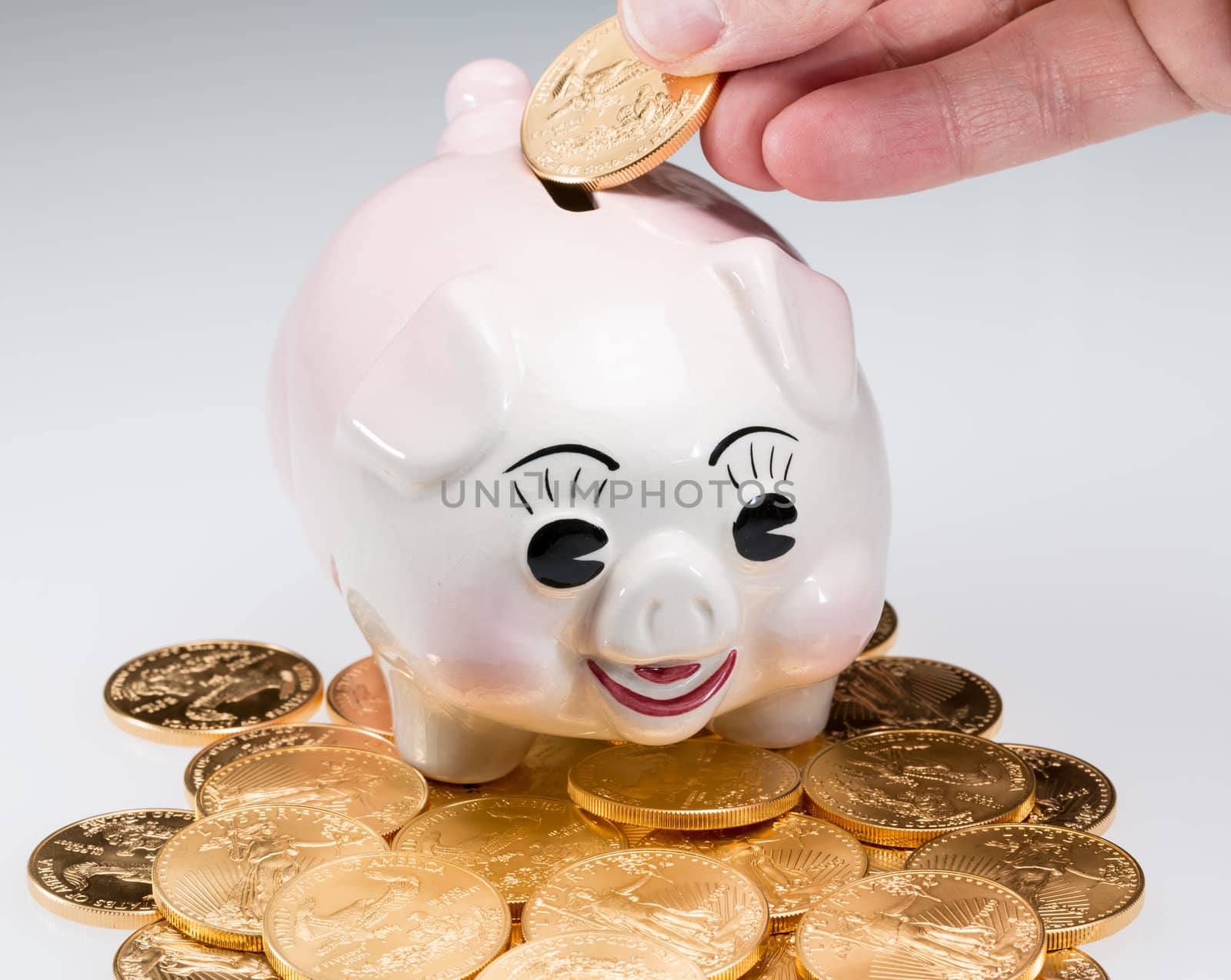 Fingers placing a single gold coin into the slot on a piggy bank with stack of coins around pig