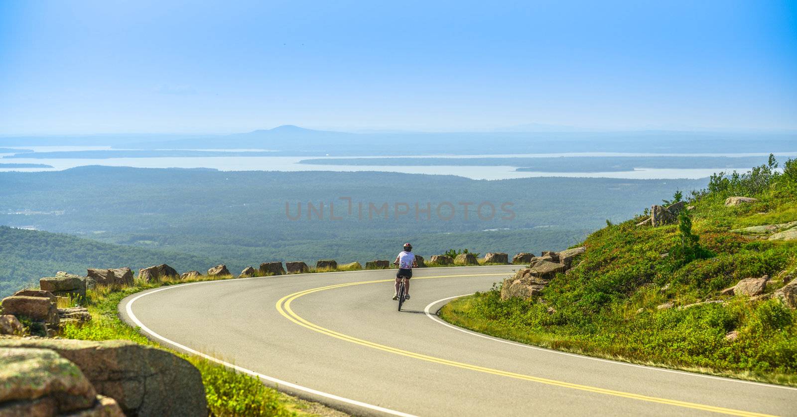Female mountain bike cyclist riding downhill along Cadillac Mountain road in Acadia National Park, a clear summer day with panormic view of Acadia National Park (Maine, USA) landscape with lakes.