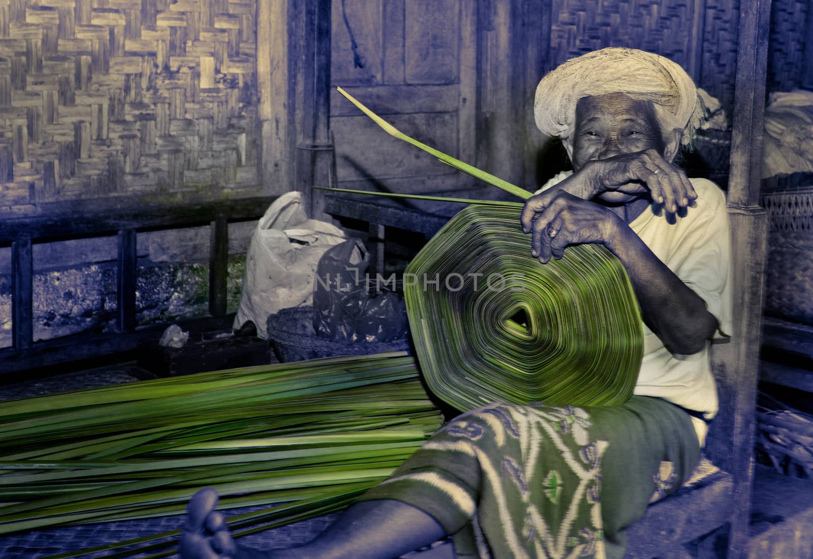 Smiling indigenous old woman sitting on a wooden bench outdoors using natural plant fibres while weaving a basket or mat in Bali