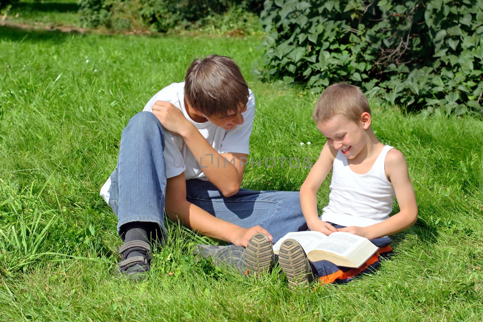 Older and Little Brothers reads a Book in the Summer Park