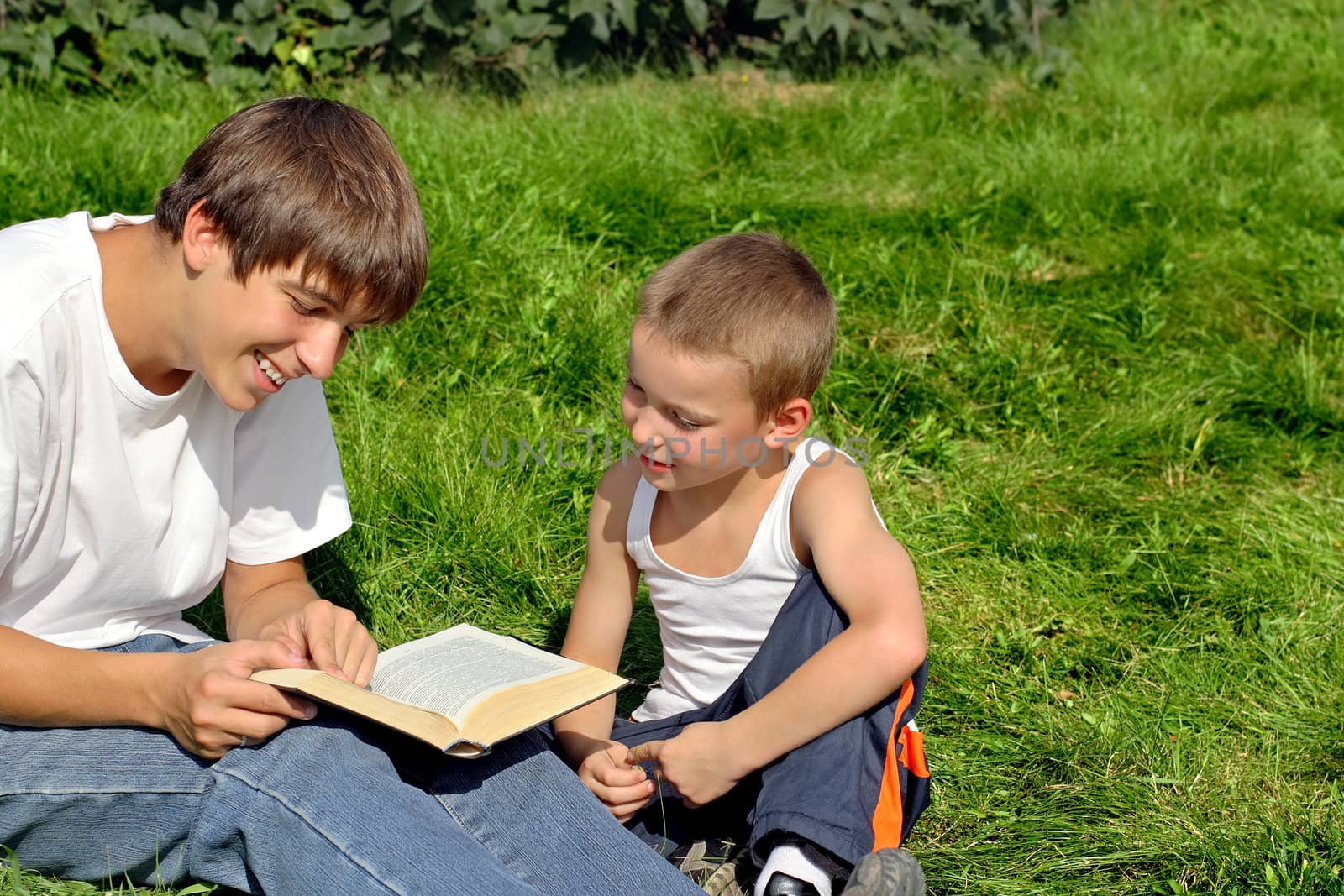 Older and Little Brothers reads a Book in the Summer Park