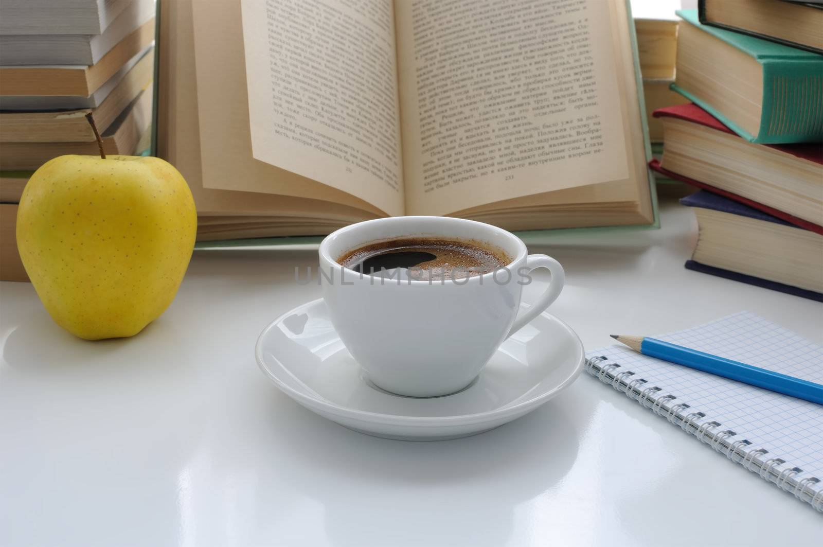A cup of coffee and an apple on a table of books with a notebook and pencil