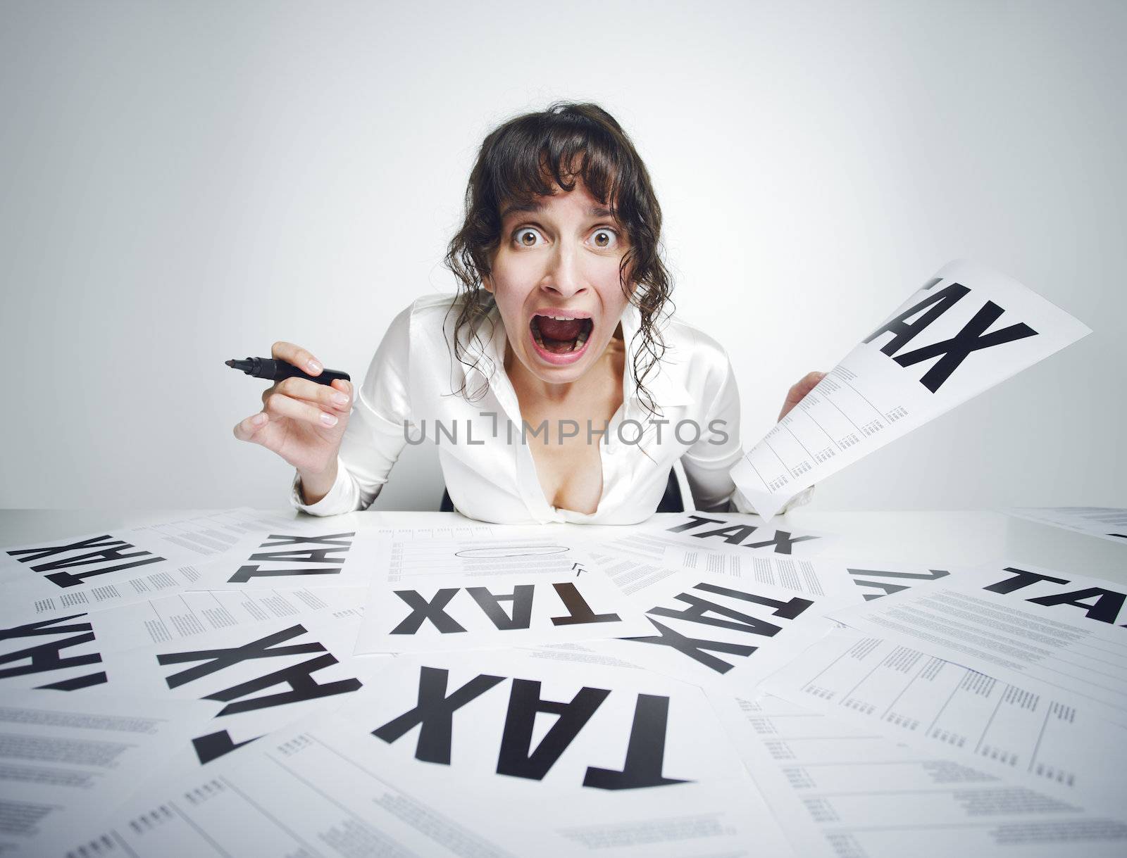 Young frightened woman shouting out while sitting at her paperwork-covered desk  with a marker in one hand and a tax bill in the other one