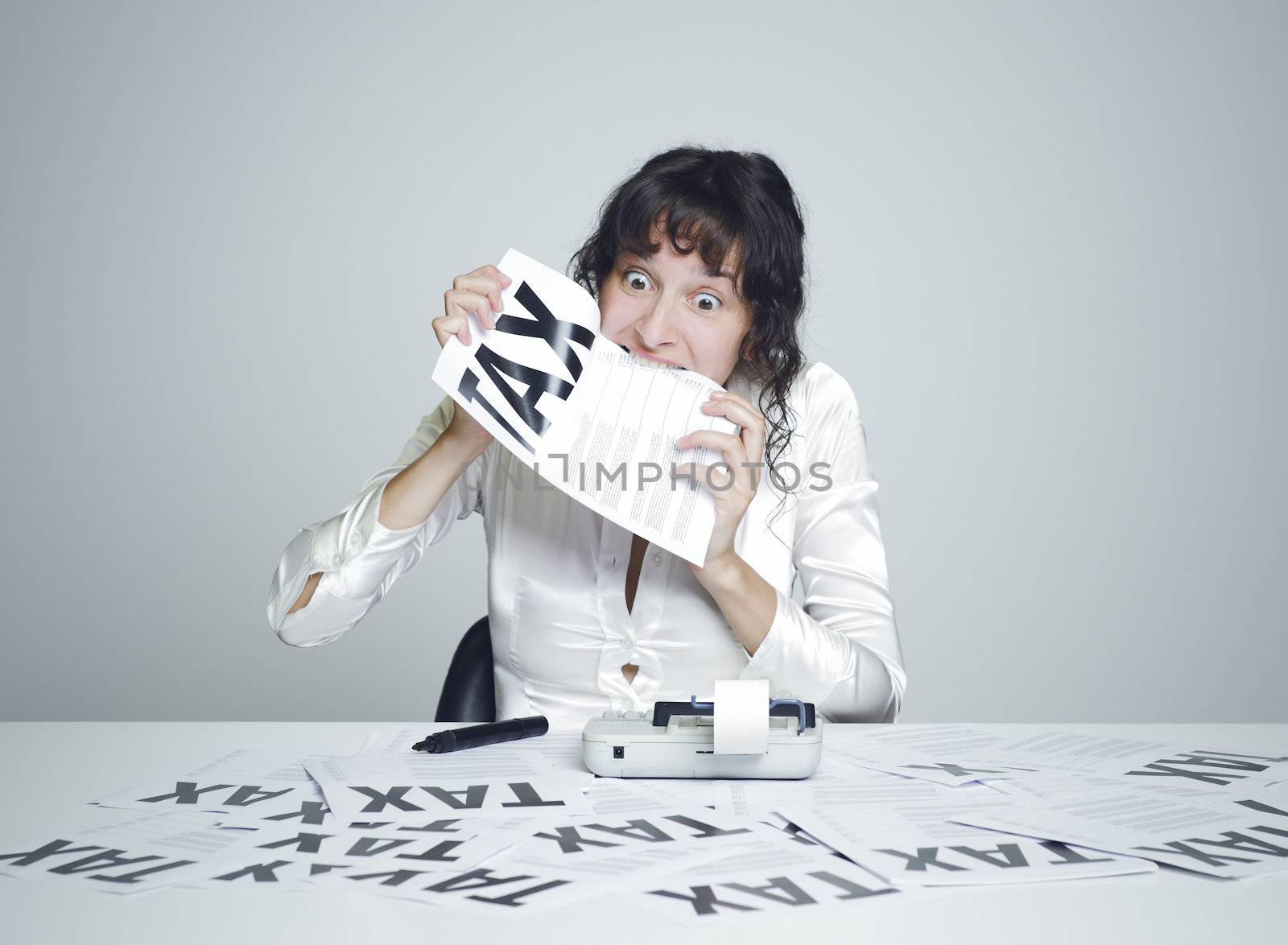 Young desperate woman at her paperwork covered desk biting a tax form