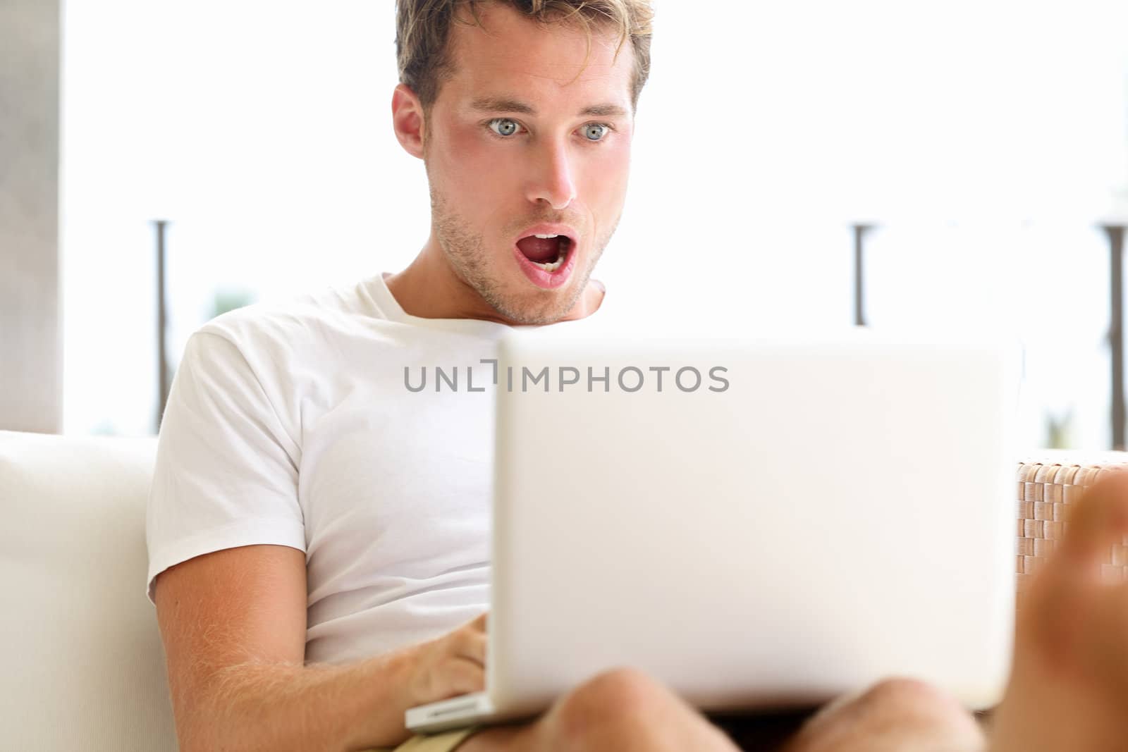 Shocked surprised man looking at laptop computer surprised and amazed with open mouth and big eyes. Funny young male model sitting outside on sofa.