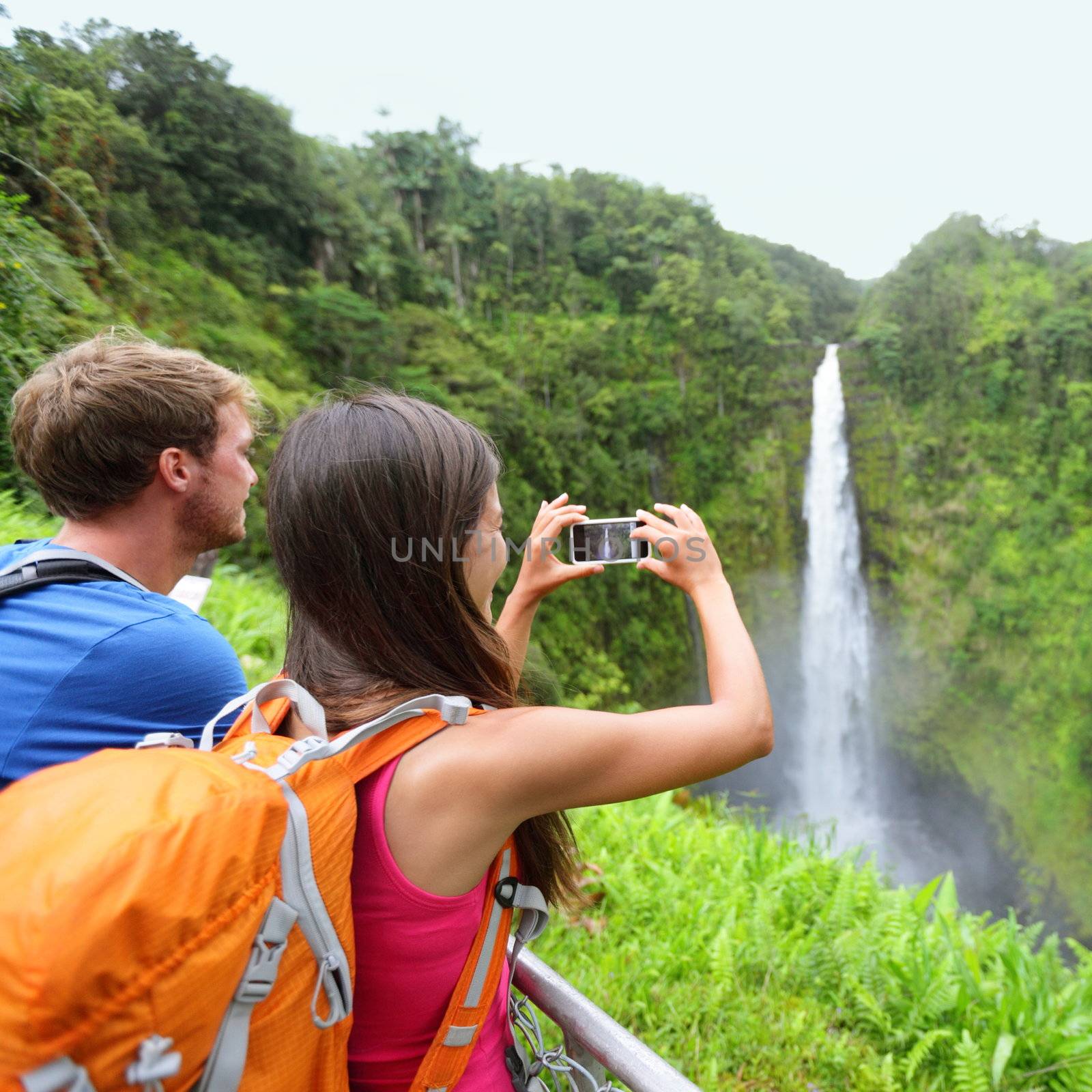 Tourist couple on Hawaii taking pictures of the famous Akaka Falls waterfall on Hawaii, Big Island, USA. Happy cheerful young multicultural couple tourists on travel.