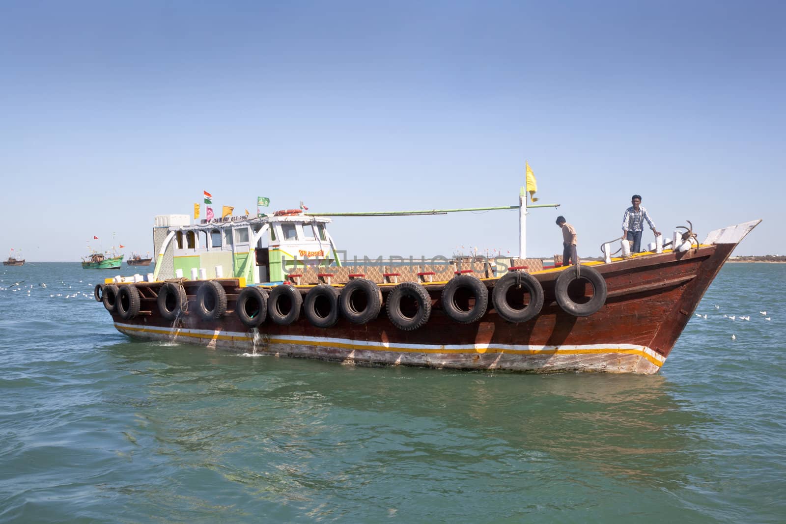 Dwarka Roadtrip. Offshore ferry boat at Dwarka with crew under a fairly clear blue sky  with other vessels in the distance and seagulls on the water surface and in flight. Gujarat India