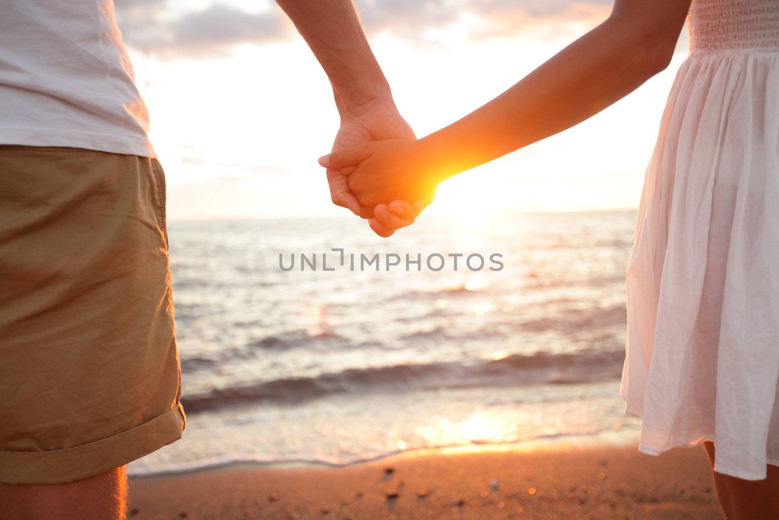 Summer couple holding hands at sunset on beach. Romantic young couple enjoying sun, sunshine, romance and love by the sea. Couple on summer vacation travel holiday.