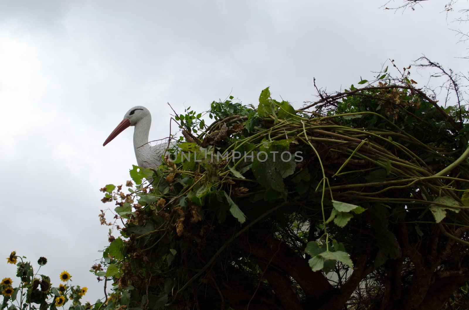 decorative plastic stork sit in handmade nest from creeper plants on background of cloudy sky.