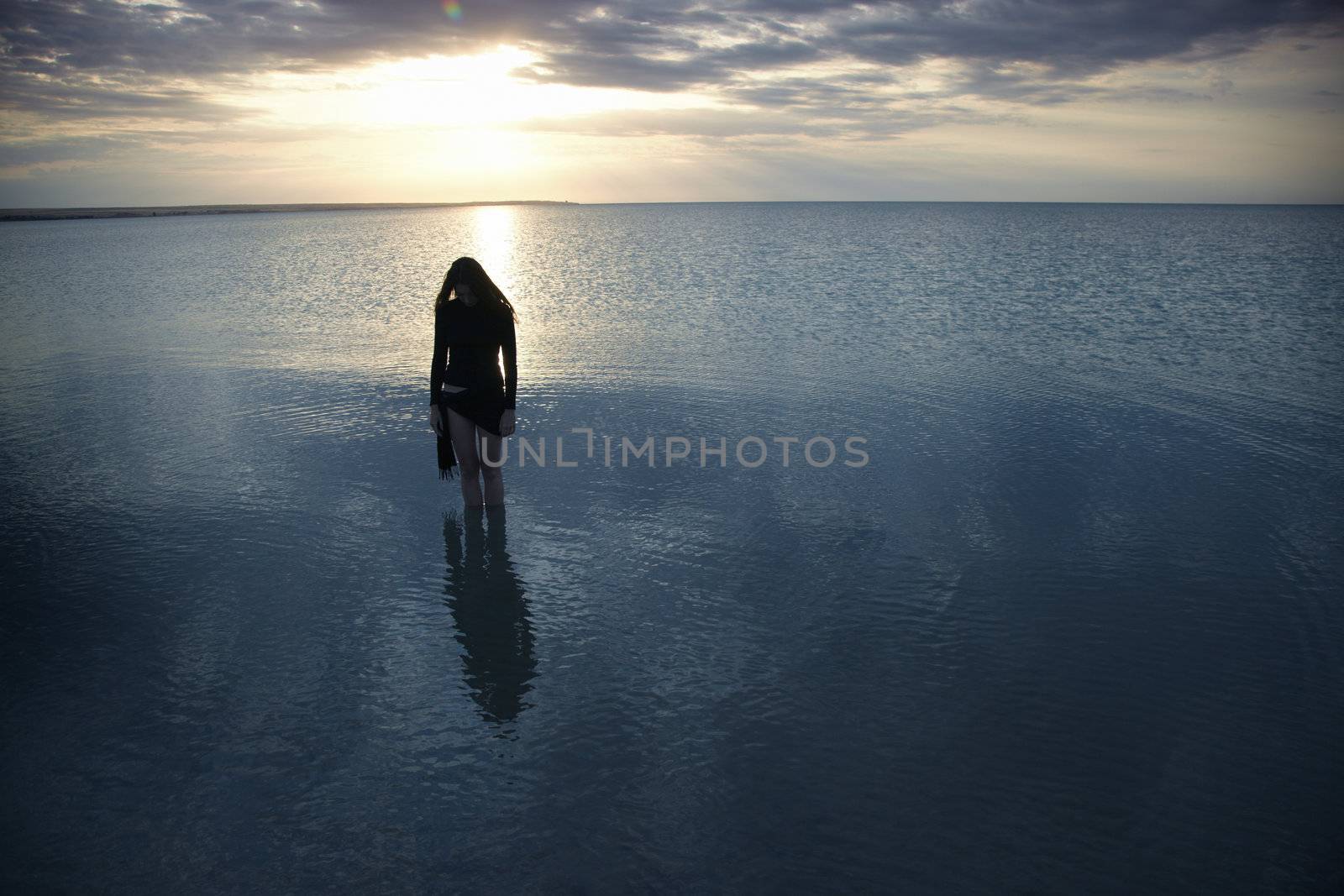 Silhouette of the single sad woman standing in the sea during sunset. Artistic colors and darkness added