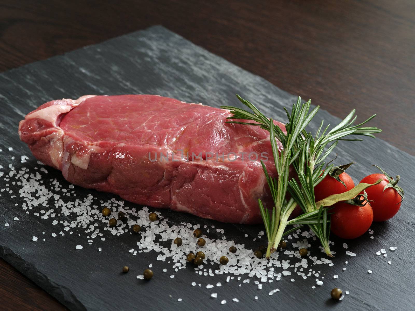 Photo of a raw thick sirloin steak with rosemary, cherry tomatoes, salt and peppercorns on a piece of black slate.