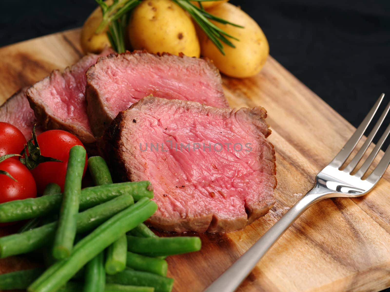 Photo of steak dinner with thick slices of sirloin, cherry tomatoes, green beans and potatoes on a wooden board.