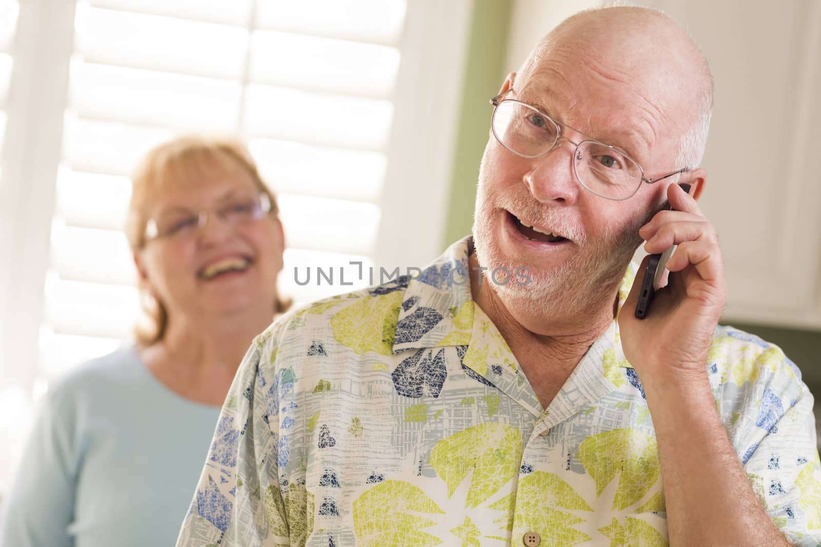 Happy Senior Adult Husband on Cell Phone with Wife Behind in Kitchen.