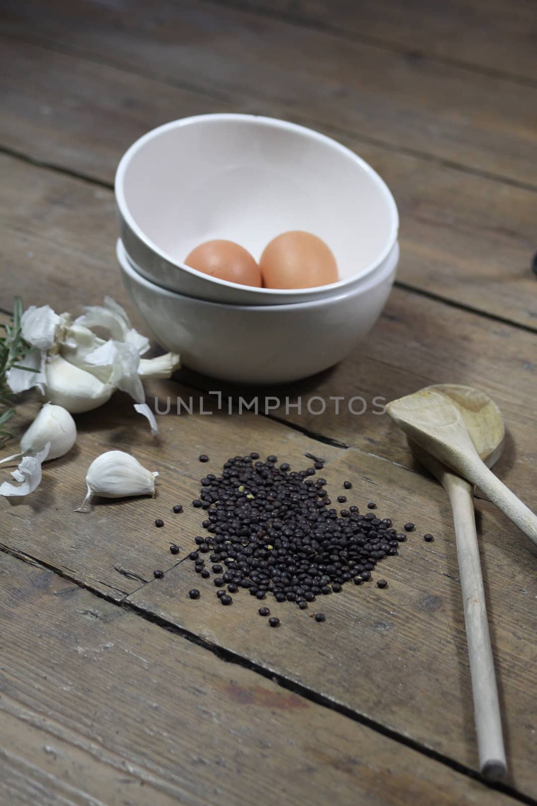 Kitchen ingredients comprising of brown eggs in a white bowl, garlic, fresh rosemary and black lentils. All set on a portrait format against a wooden background with two wooden spoons.