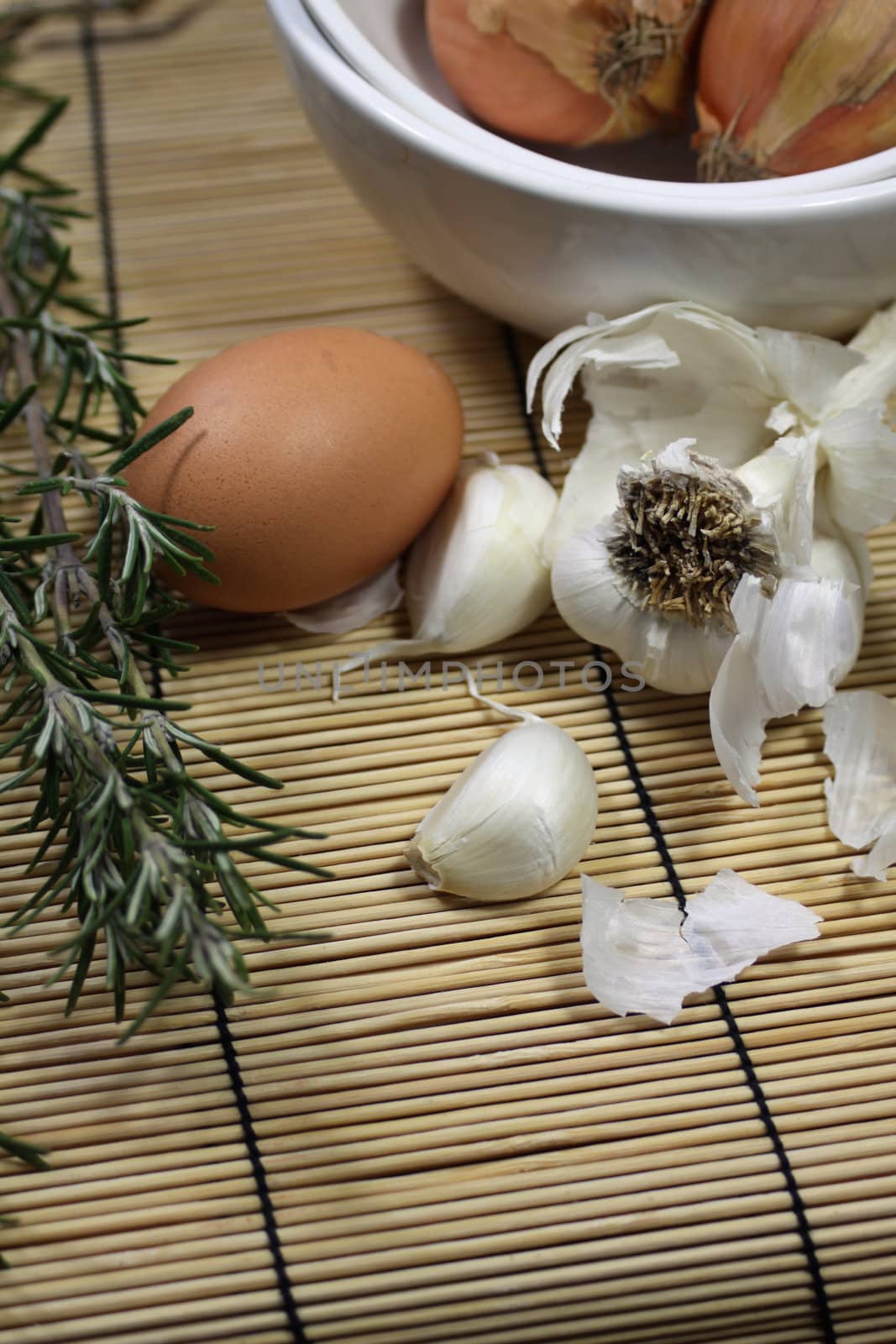 Close up detail of food ingredients comprising of a single brown egg, a bulb of garlic and a sprig of rosemary. All set on a portrait format on a bamboo matting base.