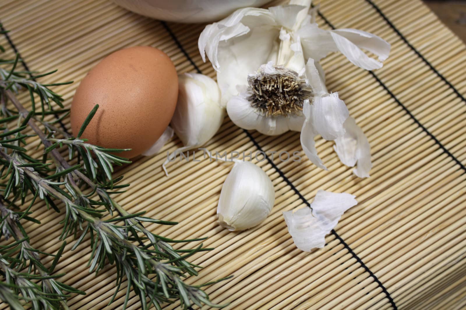 Close up detail of food ingredients comprising of a single brown egg, a bulb of garlic and a sprig of rosemary. All set on a landscape format on a bamboo matting base.