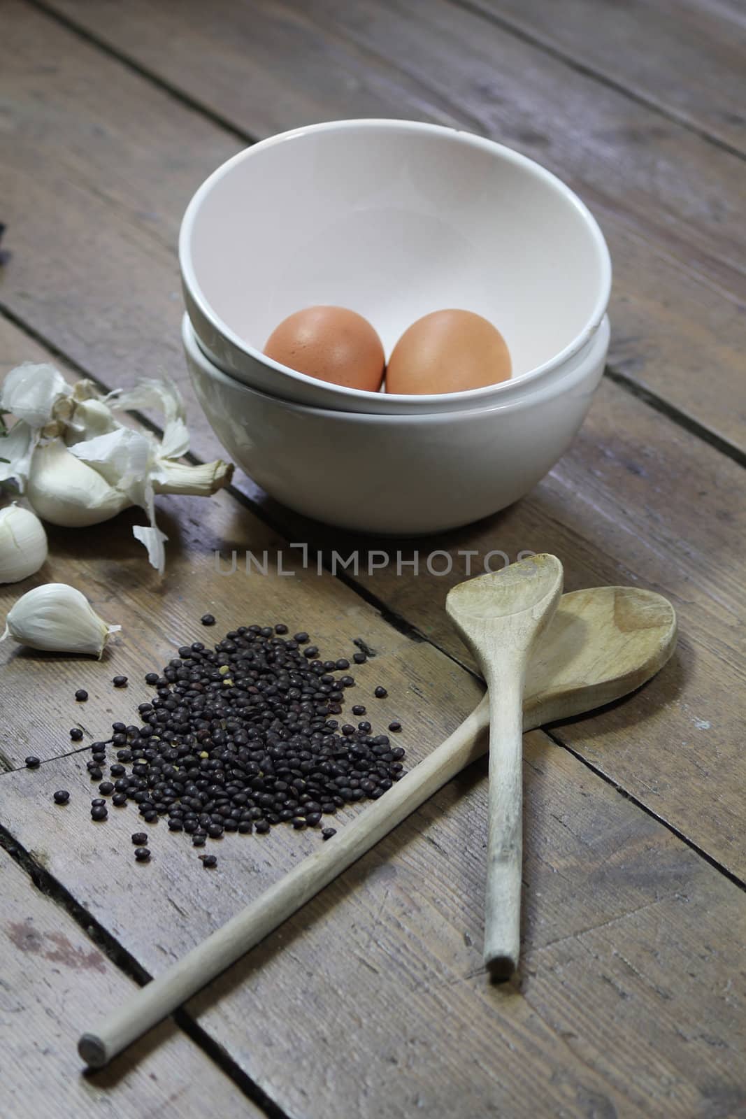 Kitchen ingredients comprising of brown eggs in a white bowl, garlic, fresh rosemary and black lentils. All set on a portrait format against a wooden background with two wooden spoons.