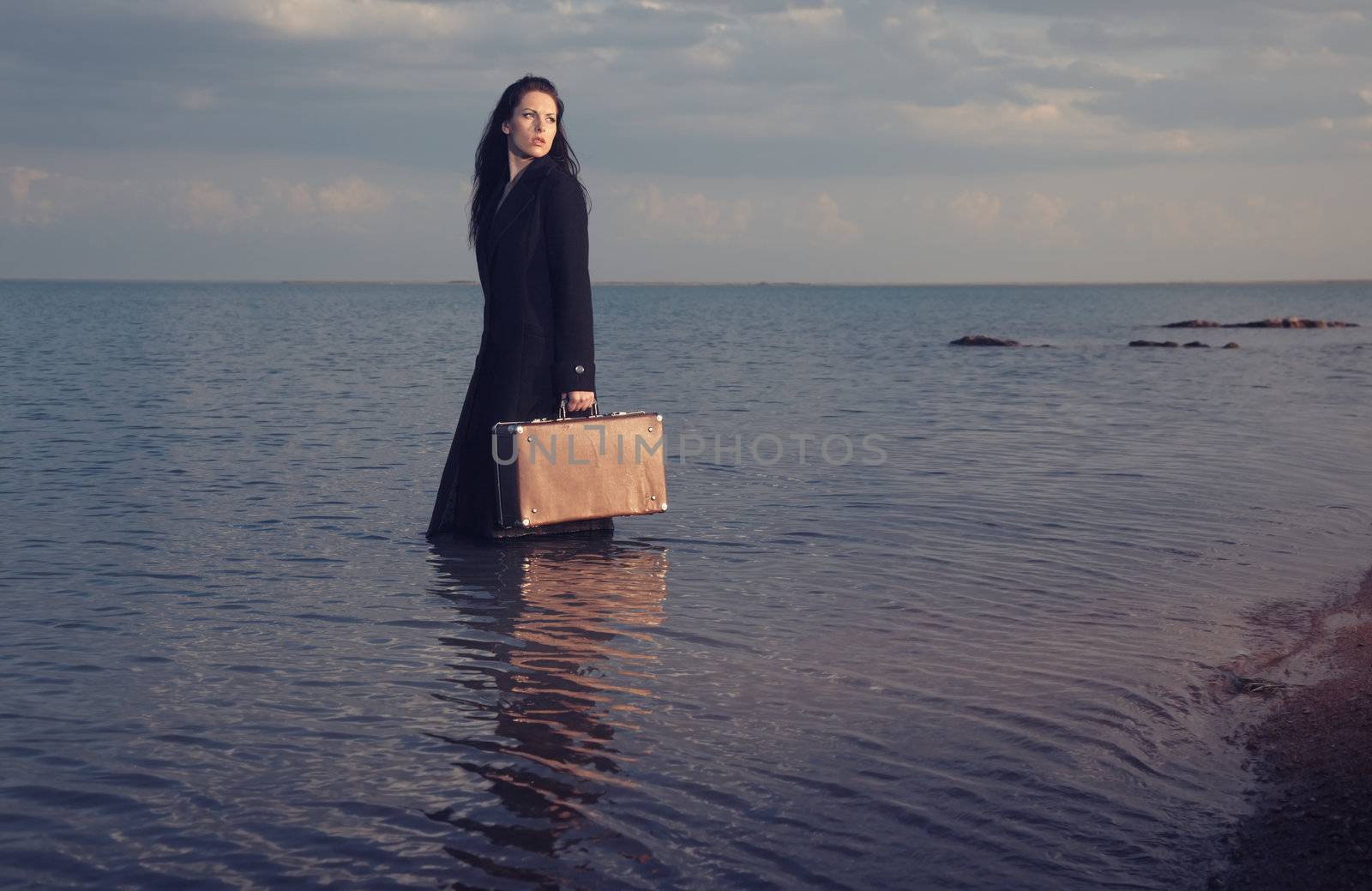 Lady in coat standing in the sea with travel bag