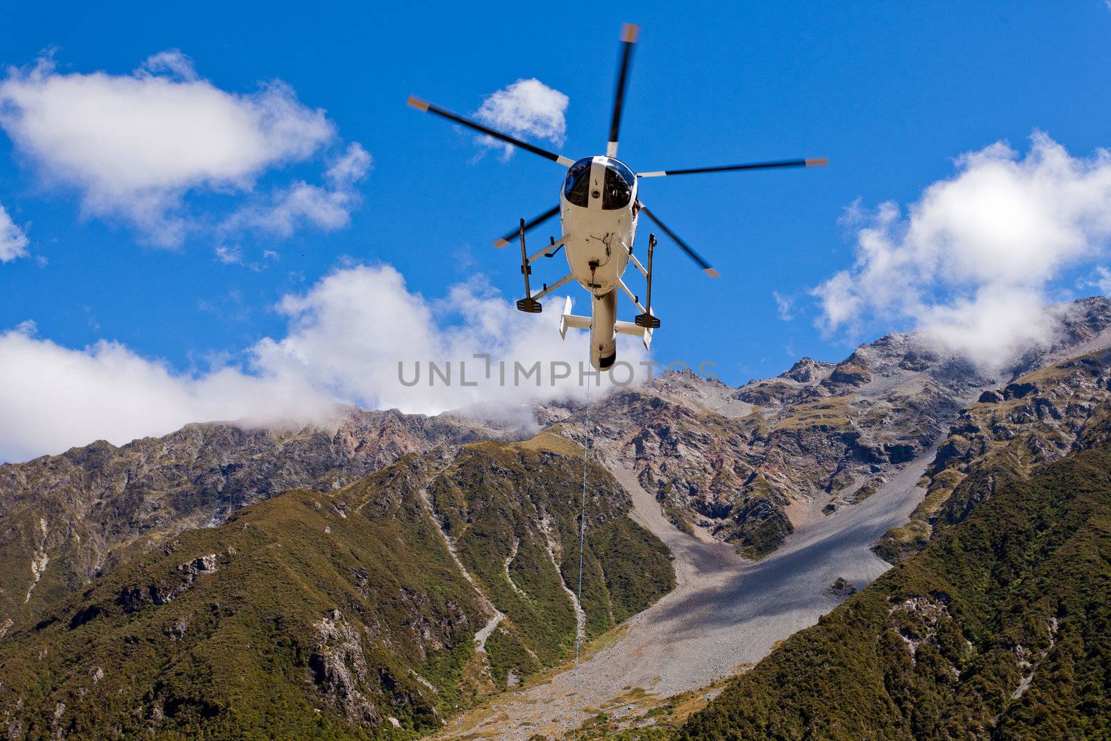 Small helicopter hovering mid-air in blue sky over mountains of Southern Alps New Zealand