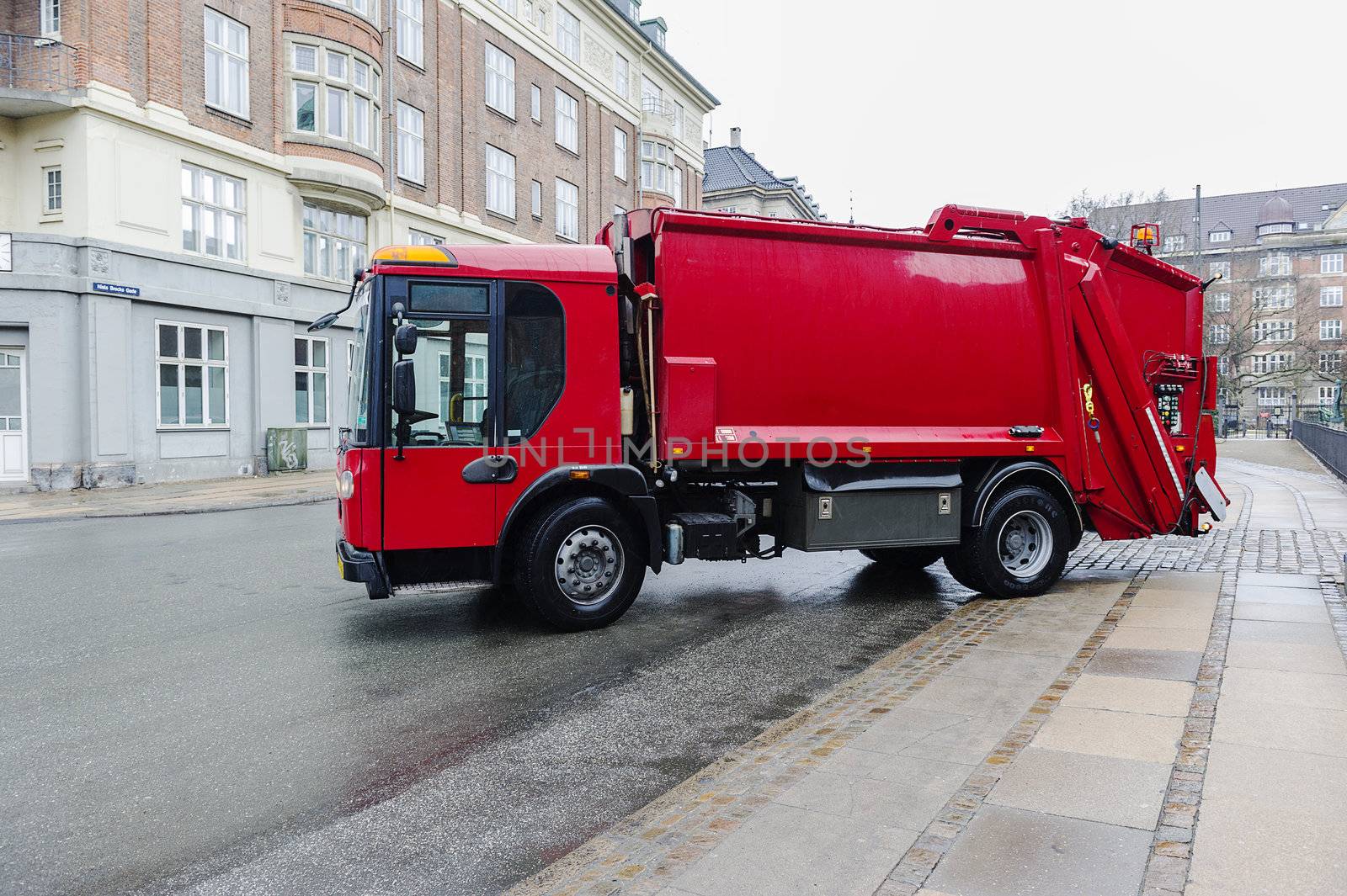 Red garbage disposal truck parked at the side of a street collecting household rubbish and waste for crushing, recycling and treatment or disposal on municipal dumps