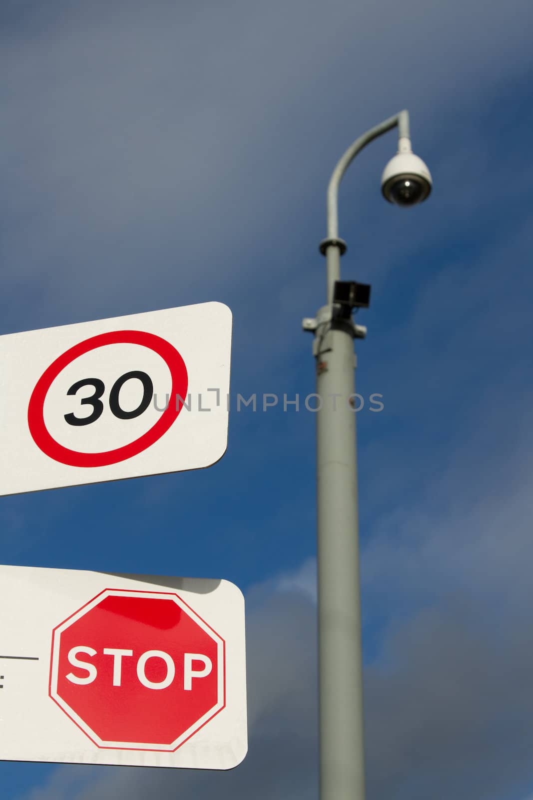 A pair of signs on a white background with a 30 speed limit and a stop warning overlooked in the background by a cctv camera.