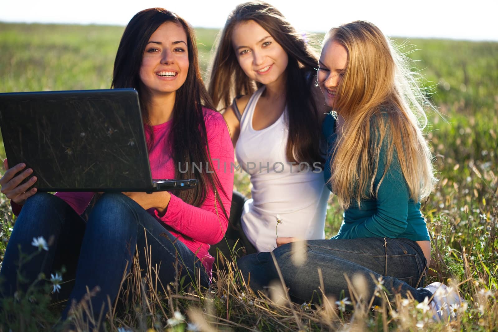 three young beautiful woman with a laptop sitting in the field o by jannyjus