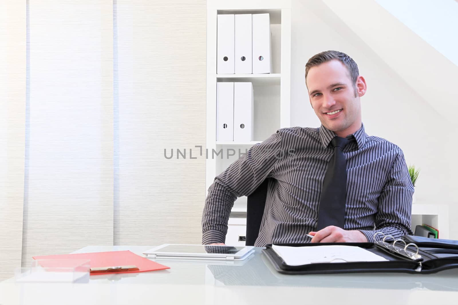 Smiling successful young businessman sitting at his desk which is spread with folders and a tablet computer