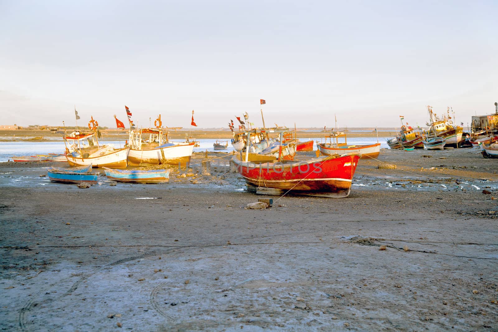 Dwarka Roadtrip. Landscape of an orange evening sun falling on banked and grounded fishing boats and ferries in Gujarat India. Hazy blue sky and silty foreground with motorbike tire marks