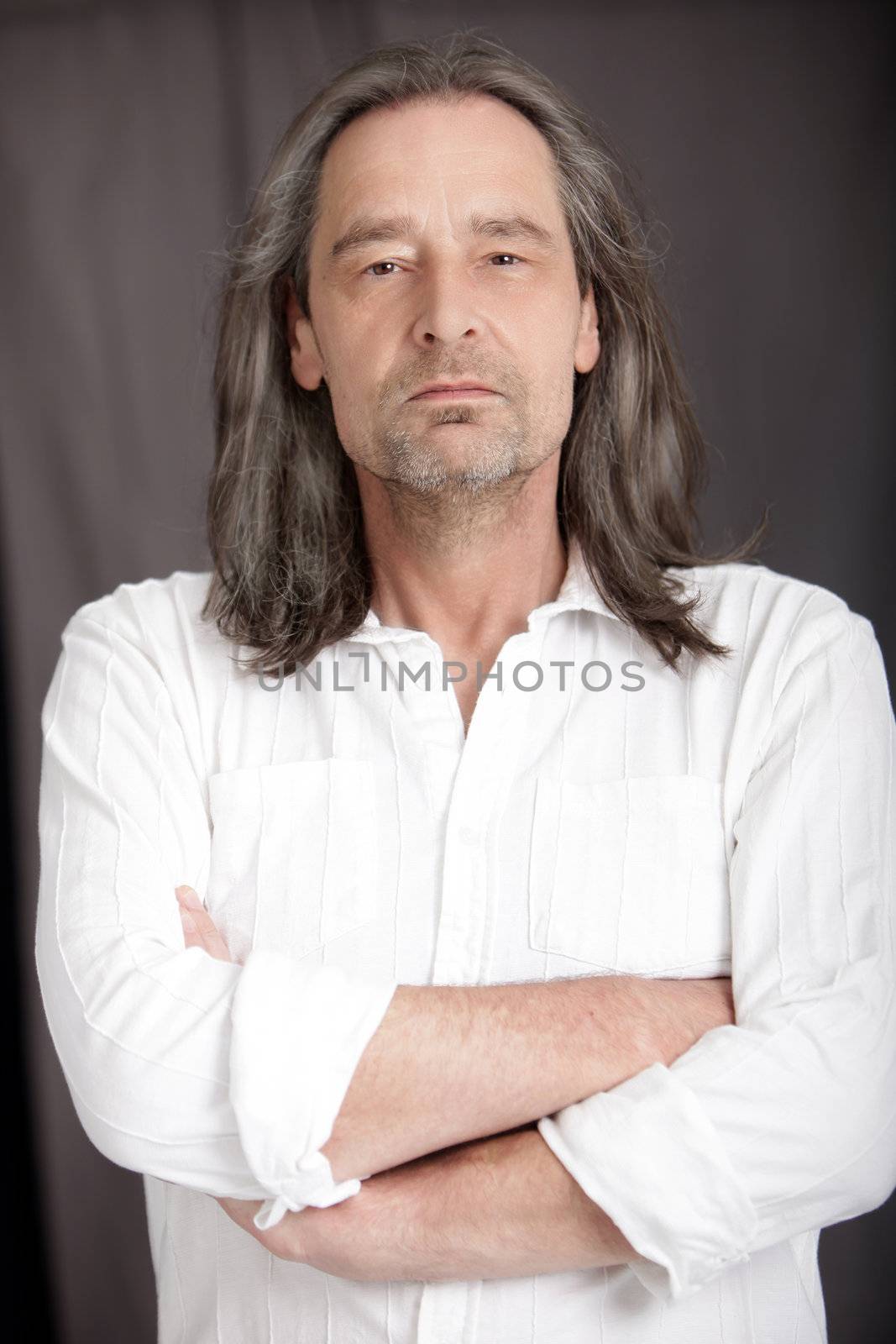 Serious middle-aged man with shoulder length hair standing with his arms folded looking at the camera, upper body studio portrait