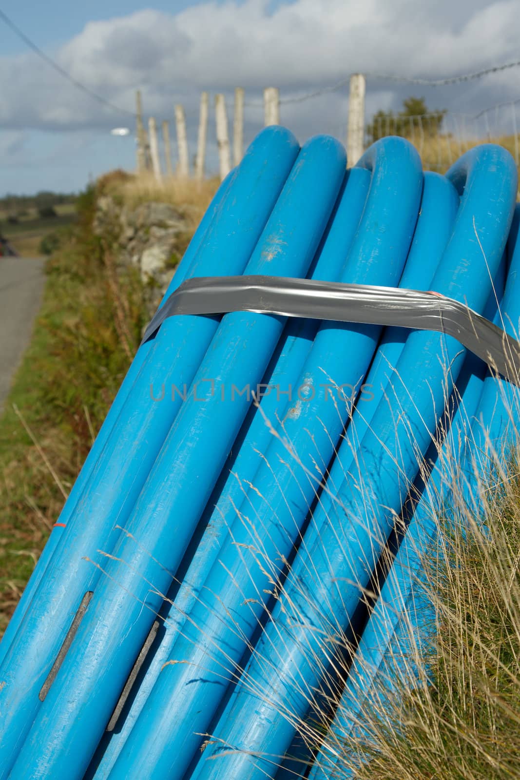 A coiled blue water pipe leaning on a wall on the side of a road verge with a fence and blue cloudy sky.