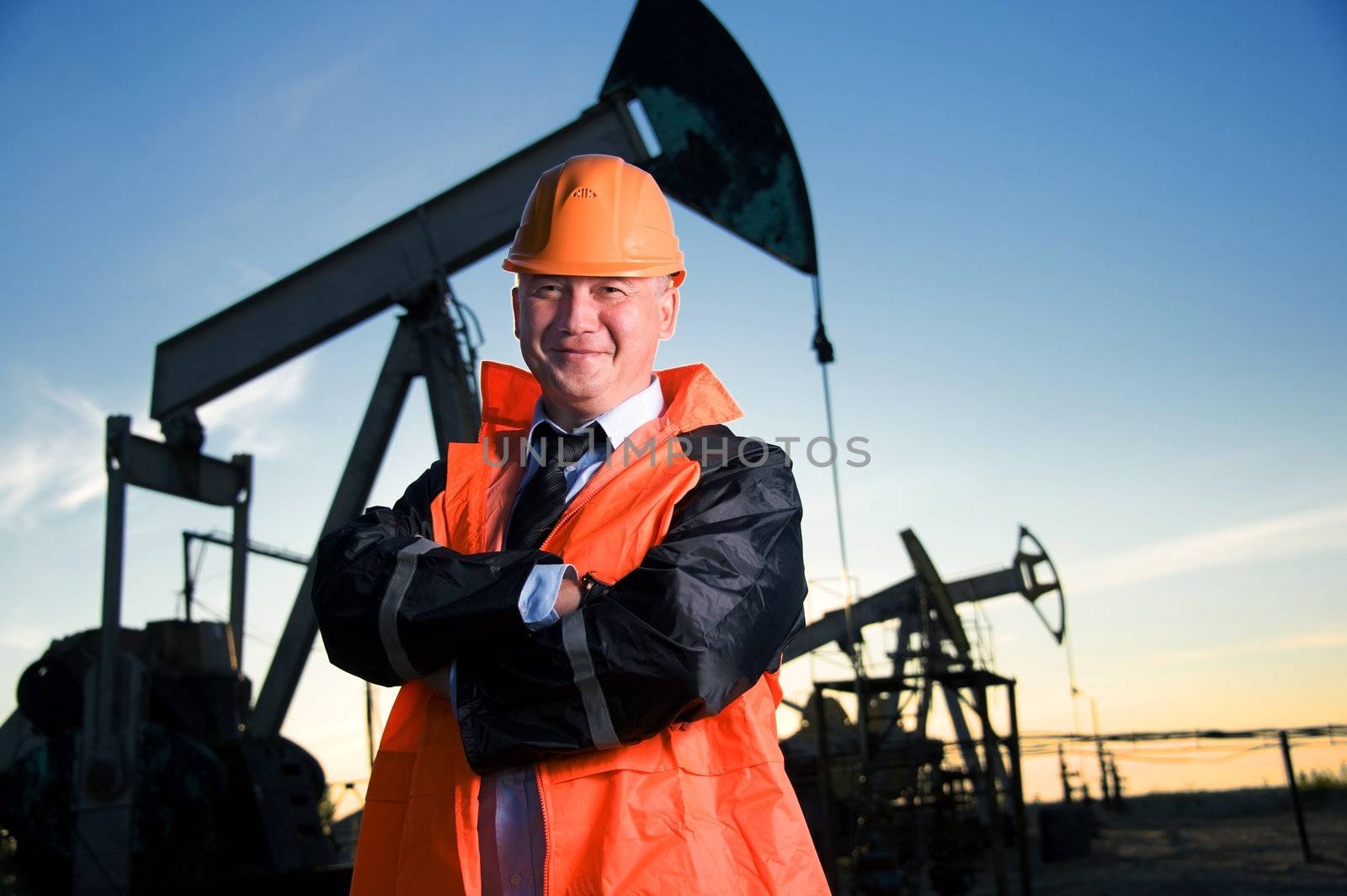 Oil worker in orange uniform and helmet on of background the pump jack and sunset sky.