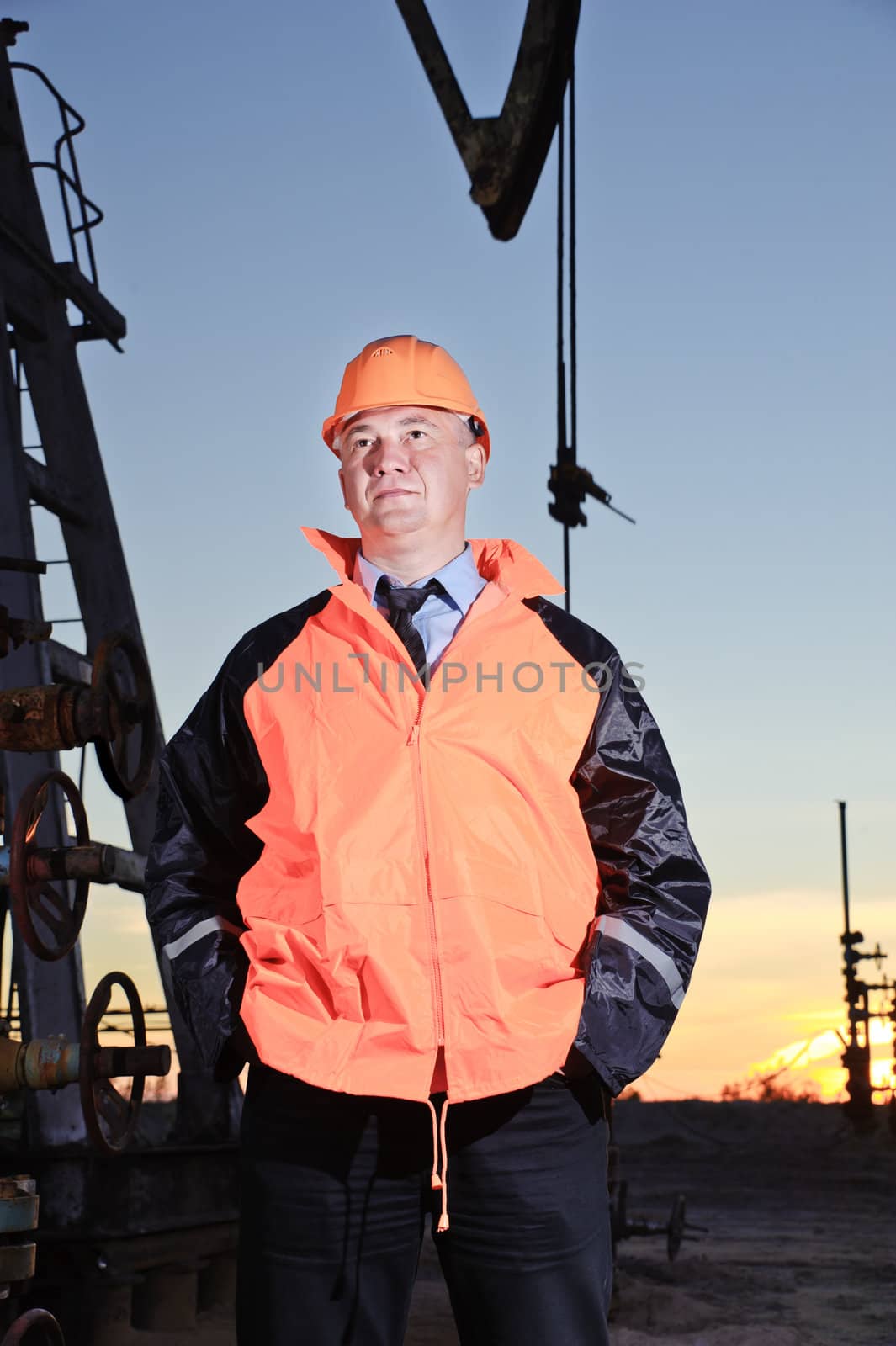 Worker in orange uniform and helmet on of background the pump jack and sunset sky. Severe. Hands in pockets.