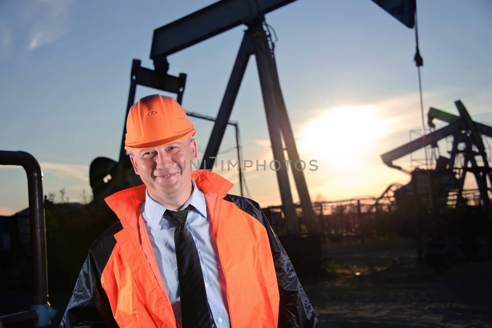 Oil worker in orange uniform and helmet on of background the pump jack and sunset sky.