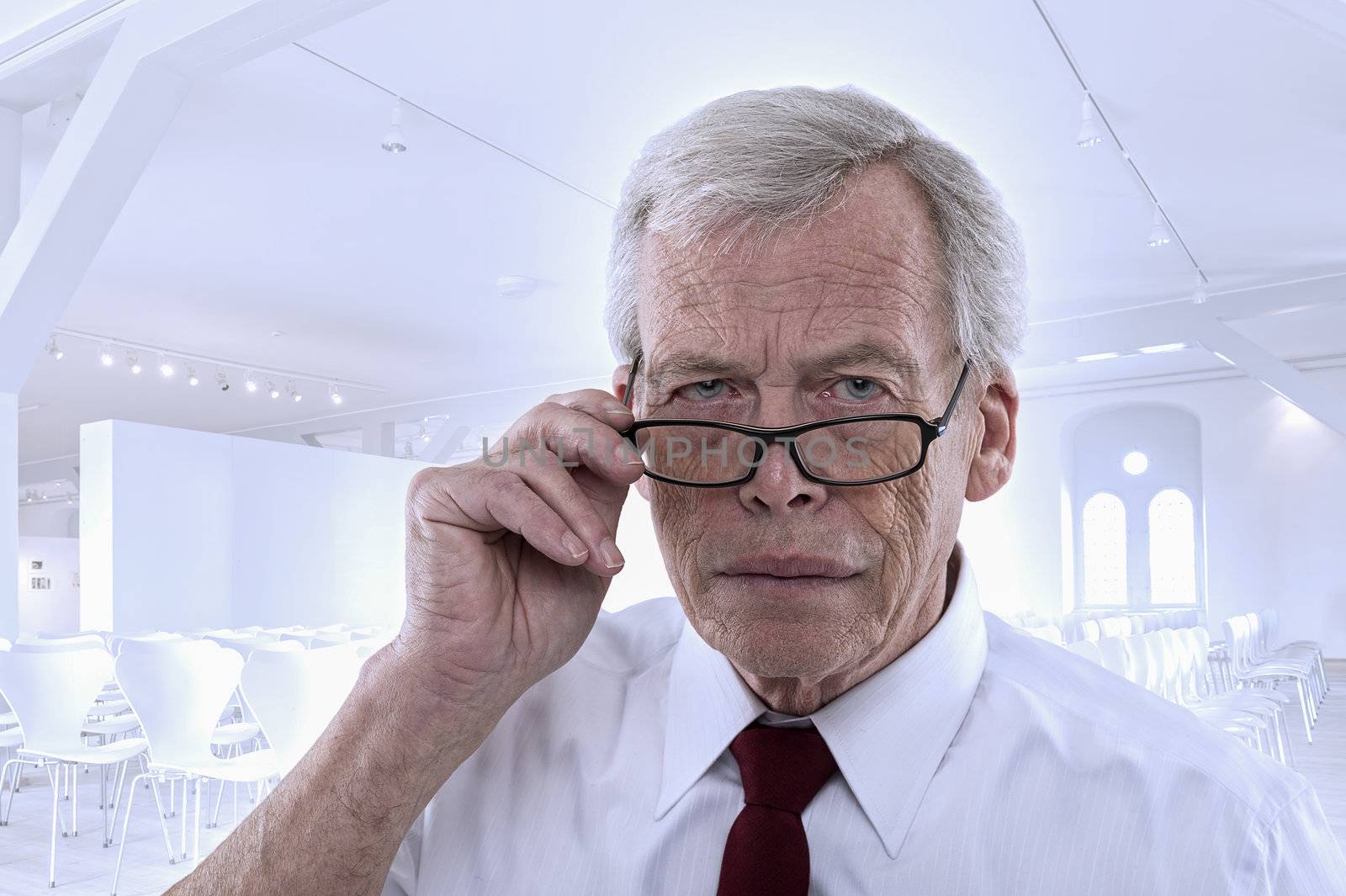 Handsome grey haired senior business man lowering his glasses and peering at the camera with a serious expression against a high key architectural background