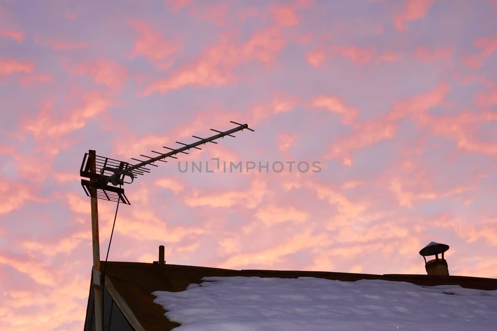 Television antenna over the snowy roof of the old house against reddish cloudy sky in evening time