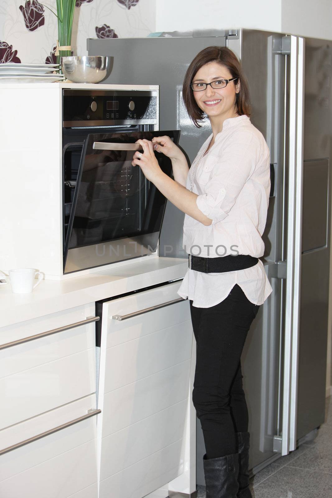 Stylish young woman cooking standing at her oven opening the door while smiling at the camera