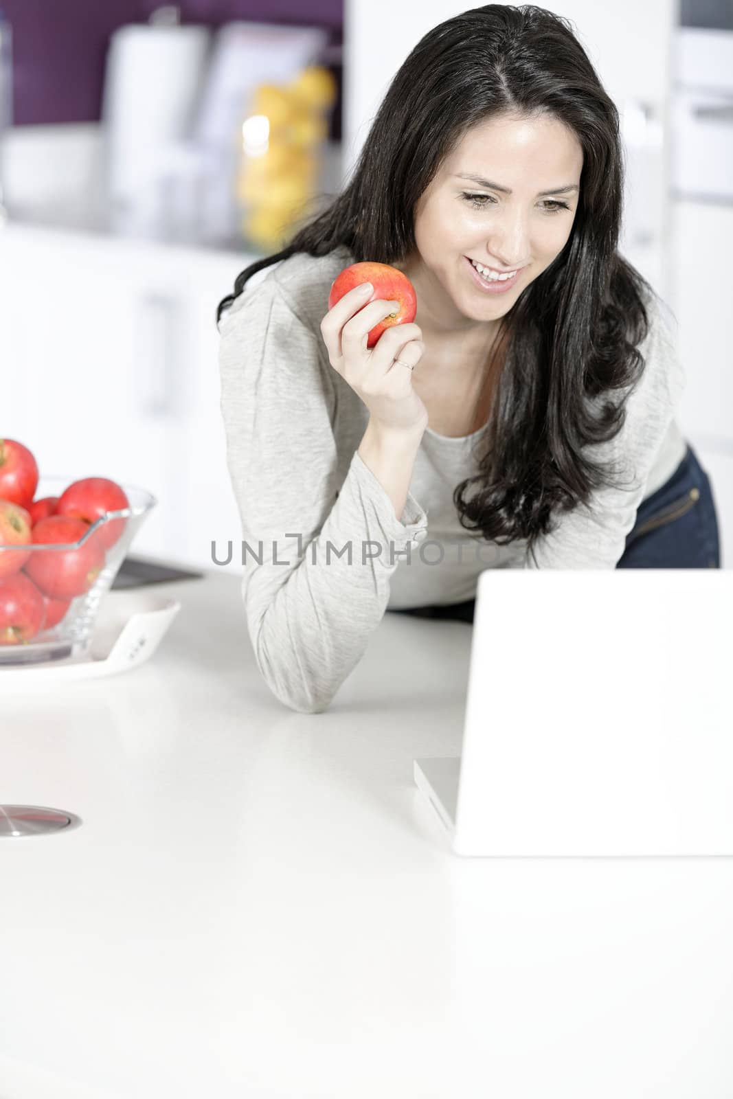 Beautiful young woman eating an apple in her white kitchen relaxing