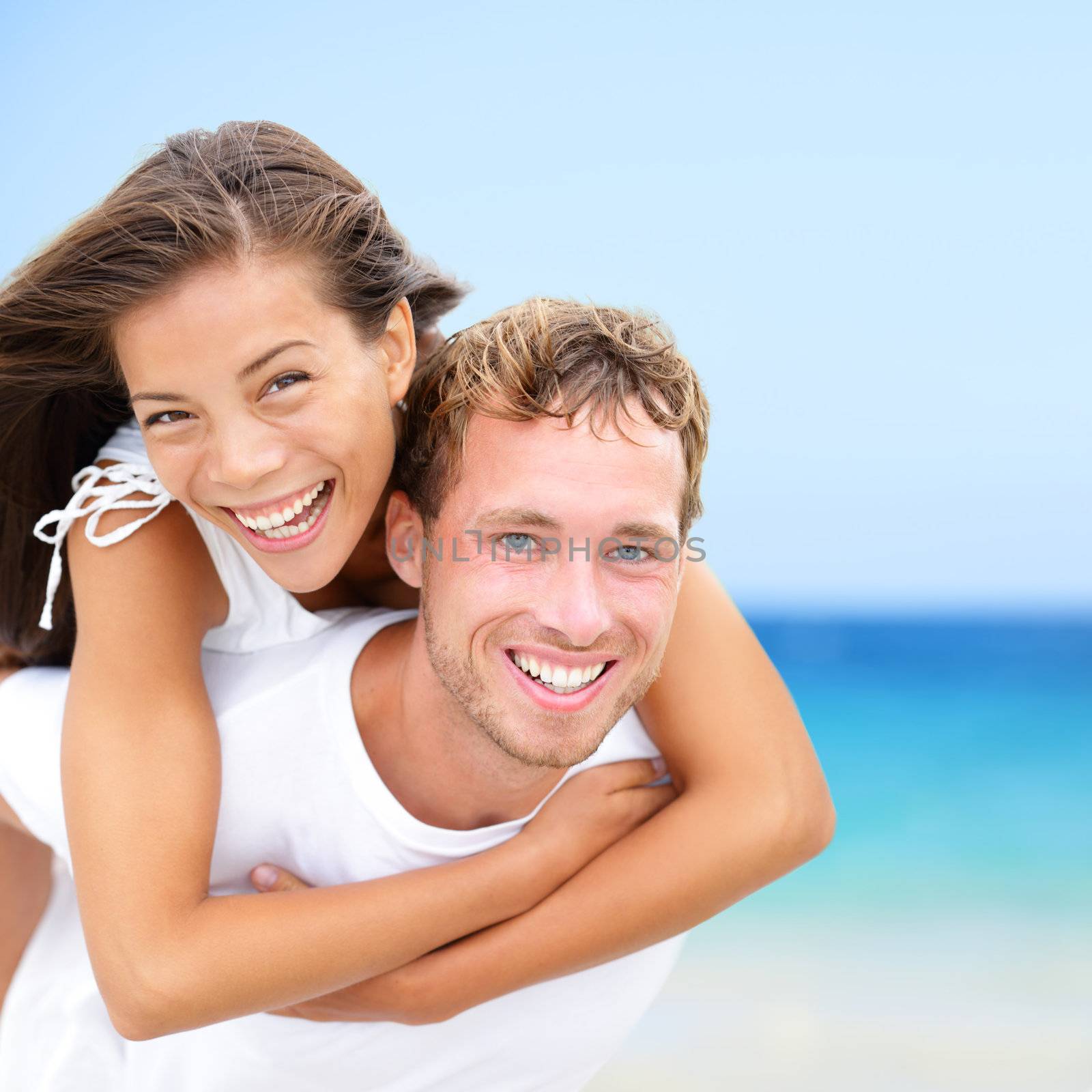 Happy couple on beach fun summer vacation. Multiracial Young newlywed couple piggybacking smiling joyful elated in happiness concept on tropical beach with blue water, sky. Asian woman, Caucasian man.